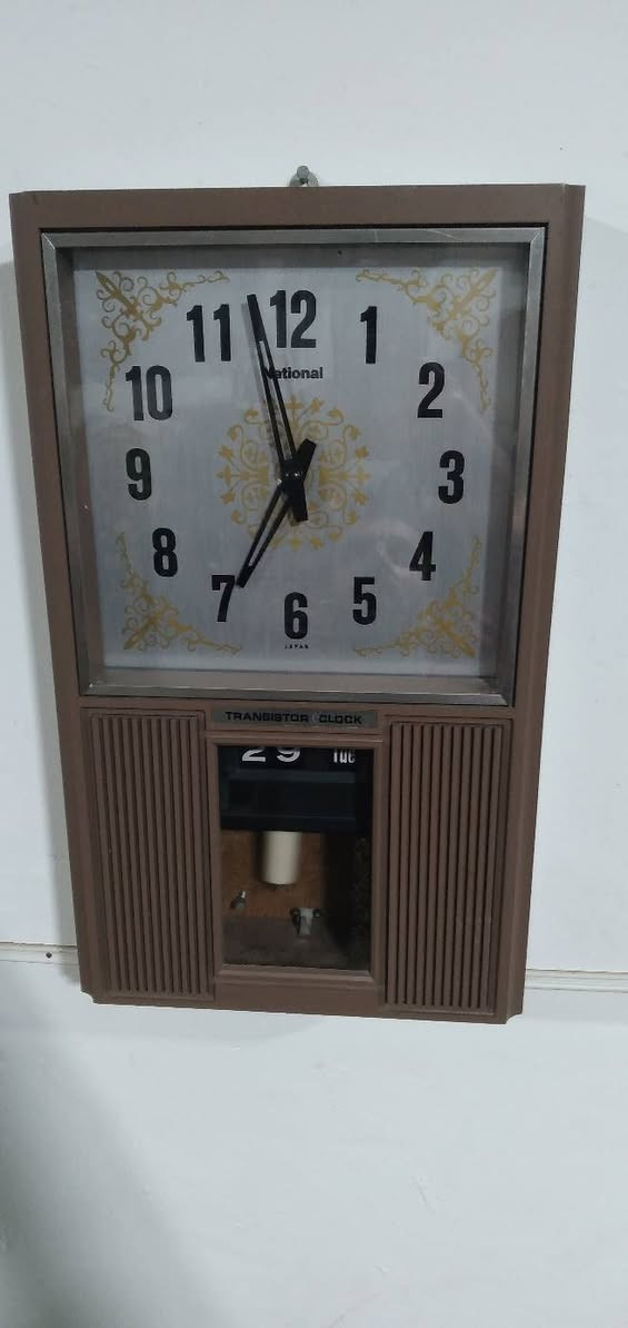 ***********
كلاسيكية رائعة جداً!  ساعة حائط يابانية عتيقة (Vintage) من إنتاج شركة National (ناشيونال - المعروفة حالياً باسم باناسونيك Panasonic)، وتحديداً من فئة ساعات "Transistor Clock" (ساعة ترانزستور) التي كانت شائعة جداً في فترة أواخر الستينيات والسبعينيات من القرن الماضي.
​الهيكل والتصميم الخارجي
​النمط المستوحى من الخمسينيات والستينيات (Mid-Century Modern): تتميز الساعة بهيكل مستطيل رأسي، مطلي باللون البني الكلاسيكي مع وجود خطوط رأسية (مشرشرة) على الجانبين السفليين تشبه فتحات المكبرات الصوتية في الراديو القديم، وهو أسلوب تصميمي كان دارجاً جداً ليعطي انطباعاً بالفخامة والدفء المنزلي.
​مينا الساعة (Dial): المينا يأتي بخلفية معدنية فضية لامعة (Brushed aluminum)، محاطة بزخارف ذهبية كلاسيكية في الأركان وفي المنتصف. الأرقام العربية كبيرة، سوداء، وواضحة جداً بخط سميك يسهل قراءته من مسافة بعيدة.
​العلامة التجارية والمنشأ:   شعار National أسفل الرقم 12، وفي الأسفل تماماً تحت الرقم 6 كُتبت عبارة JAPAN لتؤكد جودة الصناعة اليابانية في تلك الحقبة.
​ آلية العمل (الحركة والتكنولوجيا)
​Transistor Clock (ساعة الترانزستور): اللوحة الفضية الصغيرة أسفل المينا تحمل عبارة "TRANSISTOR CLOCK". هذه التكنولوجيا كانت طفرة في وقتها؛ فهي تمثل مرحلة انتقالية بين الساعات الميكانيكية بالكامل (التي تعتمد على الزنبرك والتعبئة اليدوية) وساعات الكوارتز الحديثة. تعمل هذه الساعة بالبطارية، حيث يقوم ترانزستور صغير بتنظيم النبضات الكهربائية لتحريك بندول مغناطيسي أو عجلة توازن، مما يحافظ على دقة الوقت دون الحاجة لتعبئتها يدوياً.
​البندول (Pendulum): في النافذة السفلية المحاطة بالإطار البني، يمكن رؤية جزء من البندول الداخلي الذي يتحرك يميناً ويساراً، وهو مغطى بزجاج شفاف يتيح رؤية الحركة الميكانيكية التي تعطي الساعة روحاً وجمالية خاصة.
​ الميزات الإضافية (التقويم)
​نافذة التاريخ واليوم (Day/Date Calendar): بجانب البندول في القسم السفلي، تحتوي الساعة على تقويم ميكانيكي يظهر اليوم والشهر (يظهر في الصورة رقم 29 واليوم "Tue" أي الثلاثاء). هذا النوع من التقويم يتغير تلقائياً مع حركة عقارب الساعة عند منتصف الليل.
​باختصار، هذه الساعة ليست مجرد أداة لمعرفة الوقت، بل هي قطعة فنية تجسد ذروة الصناعة الهندسية والجمالية اليابانية في السبعينيات، وتعتبر اليوم من القطع المميزة جداً لهواة جمع المقتنيات والأنتيكات الإلكترو-ميكانيكية.
