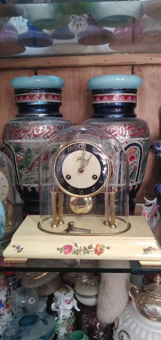 ساعة مكتبية أو رفّية (Mantel/Table Clock) كلاسيكية مميزة.  
​العلامة التجارية:   اسم الساعه "Selva"، وهي علامة تجارية ألمانية معروفة جداً في مجال قطع غيار الساعات، ومجموعات تركيب الساعات، والساعات الميكانيكية التقليدية بجودة عالية.
​المنشأ:  أسفل الرقم 6 عبارة صغيرة  مكتوب "Made in Germany" (صنع في ألمانيا)، وهو أمر متوافق تماماً مع العلامة التجارية Selva.
​ التصميم والهيكل
​القبة الزجاجية (Glass Dome): الساعة مغطاة بقبة زجاجية شفافة منحنية (تُعرف بساعات القبة أو ساعات الذكرى السنوية أحياناً، وإن كانت هذه تعمل ببندول تقليدي وليس بندولاً دواراً). وظيفتها حماية الآلية الداخلية والميناء من الأتربة.
​الميناء (Dial): يتميز بتصميم ثنائي اللون (حلقة خارجية سوداء تحتوي على أرقام عربية مذهبة واضحة، ومركز دائري بلون كريمي/أبيض مطفأ).
​الأعمدة والقاعدة: ترتكز الساعة على أعمدة نحاسية مضلعة مذهبة فوق قاعدة بيضاء أو كريمية المزينة بنقوش وردية ناعمة مرسومة باليد، مما يعطيها طابعاً أنثوياً أو ريفياً كلاسيكياً (Shabby Chic / Vintage style).
​الآلية والتشغيل (الميكانيكا)
​المفاتيح وثقوب التعبئة: يحتوي الميناء على ثقبين للتعبئة (Winding holes) بجانب الرقمين 4 و 8. هذا يعني أن الساعة تحتوي على نابضين (زُنبركين): واحد لتشغيل حركة عقارب الساعة، والآخر مخصص لآلية الرنين (الدق أو الجرس).
​مفتاح التعبئة:  مفتاح التعبئة المعدني الأصلي مستلقياً على قاعدة الساعة داخل القبة الزجاجية.
​البندول (Pendulum): بندول نحاسي لامع أسفل الميناء، وهو المسؤول عن تنظيم الوقت عبر حركته الاهتزازية يميناً ويساراً.
​ الحالة العامة  الساعة في حالة جيدة جداً ونظيفة. النحاس يحتفظ بلمعانه، والزجاج سليم لا تظهر فيه شروخ واضحة، والرسومات على القاعدة بحالة ممتازة.
​القيمة: هذا النوع من الساعات يجمع بين القيمة الوظيفية والجمالية كقطع ديكور "أنتيك" أو عتيقة مرغوبة لمحبي الساعات الألمانية الميكانيكية.


**إذا كنت صاحب هذا الإعلان وتريد حذفه لأي سبب، رجاءا أرسل رسالة إلى الدعم الفني**