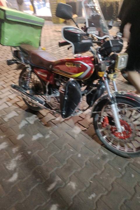 دراجة نارية للبيع، لونها أحمر مع تصميم رياضي وجذاب. تتضمن صندوق أخضر خلفي عملي لحفظ الأغراض. مثالية للتنقل في المدينة أو التوصيل. الواجه الأمامية مزودة بحاجب شمس لحماية العينين أثناء القيادة. مناسبة لمن يبحث عن وسيلة نقل اقتصادية وسهلة التعامل.