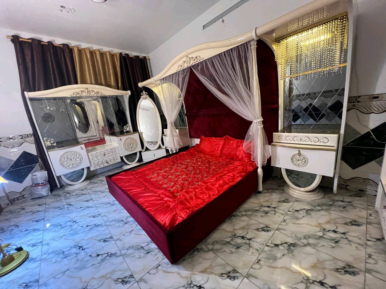 غرفة نوم تركية بتصميم فاخر وجميل، تتكون من سرير مزود بستارة شبكية وستائر مضاءة، بالإضافة إلى خزانة كبيرة مع مرايا. التصميم يجمع بين اللونين الأبيض والذهبي مع تغطية السرير بلون أحمر فاقع، مناسب لإضافة لمسة من الأناقة والرفاهية لغرفتك.