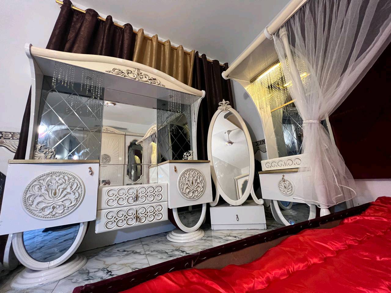 غرفة نوم تركية بتصميم فاخر وجميل، تتكون من سرير مزود بستارة شبكية وستائر مضاءة، بالإضافة إلى خزانة كبيرة مع مرايا. التصميم يجمع بين اللونين الأبيض والذهبي مع تغطية السرير بلون أحمر فاقع، مناسب لإضافة لمسة من الأناقة والرفاهية لغرفتك.