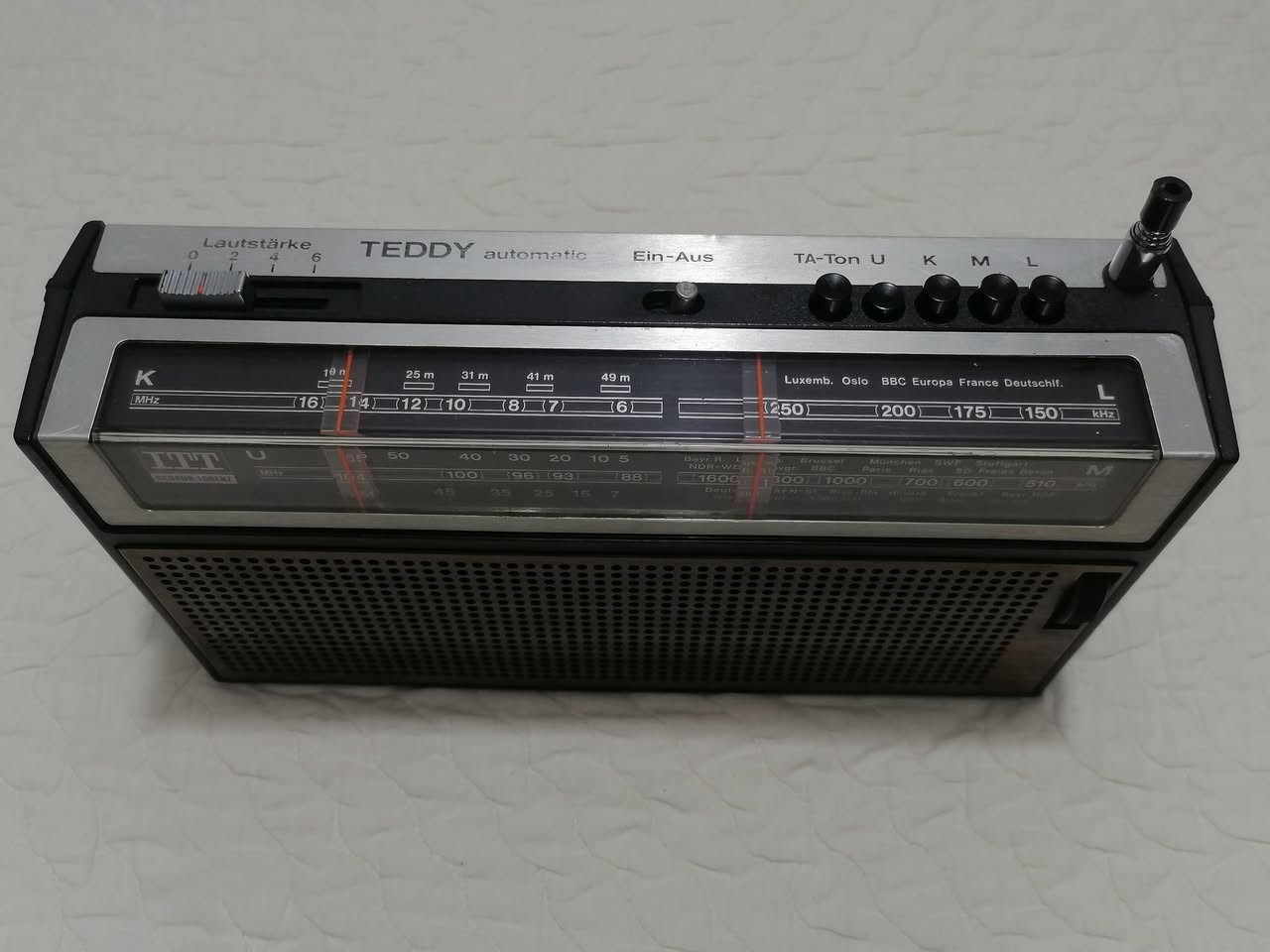 هذا الجهاز هو راديو ترانزستور كلاسيكي من نوع ITT Schaub-Lorenz Teddy automatic 103، وهو إصدار ألماني الصنع يعود لفترة أوائل السبعينيات (تحديداً بين عامي 1972 و1974).
يُعرف هذا الموديل بتصميمه العتيق (Vintage) الذي يجمع بين البلاستيك واللمسات الخشبية على الواجهة الأمامية. 
المواصفات التقنية الرئيسية:
نطاقات الموجات: يدعم استقبال موجات FM (أو UKW)، والموجات المتوسطة (MW)، والطويلة (LW)، والقصيرة (SW).
الطاقة: يعمل بالكهرباء المباشرة (110-240 فولت) أو عن طريق البطاريات (4 بطاريات وسط بقوة 1.5 فولت من نوع AA).
المكونات الداخلية: يحتوي على 11 ترانزستور، ويتميز بوجود هوائي تلسكوبي بطول 80 سم لاستقبال أفضل للموجات.
المنافذ: مزود بمقبس لسماعة الرأس (3.5 ملم) ومنفذ DIN خماسي لتوصيل مسجل صوت أو مشغل أقراص.
الأبعاد والوزن: يبلغ وزنه حوالي 1.2 كجم، وبأبعاد تقريبية 215 × 127 × 70 ملم. 
يُعتبر هذا الراديو قطعة "أنتيك" مميزة لهواة جمع الأجهزة القديمة، وغالباً ما يتم تداوله في أسواق التحفيات المستعملة .
العنوان بغداد - البياع موجود ***********‪ واتساب
