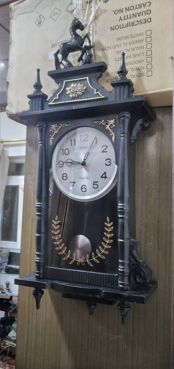 ساعة حائط بـ بندول (رقاص) من إنتاج شركة Rhythm اليابانية، وهي شركة عريقة معروفة بدقة محركاتها وتصاميمها الكلاسيكية.
​
​ هيكل الخارجي
​طراز: كلاسيكي مستوحى من طراز "منظم فيينا" (Vienna Regulator)، والذي يتميز بشكل طولي مع واجهة زجاجية تسمح برؤية البندول.
​اللون: الهيكل أسود المطفي مع لمسات تزيينية باللون الذهبي.
​ يوجد تمثال لحصان واثق، وهو عنصر تزييني تقليدي يمنح الساعة مظهراً فخماً.
​الأعمدة: على جانبي الساعة توجد أعمدة محفورة تعطي طابعاً معمارياً قديماً.
​النقوش: توجد زخارف نباتية (أزهار) في الجزء العلوي، وإكليل من الأوراق الذهبية على الزجاج السفلي يحيط بمجال حركة البندول.
​العلامة التجارية:  Rhythm   الموديل الذي يتميز بنغمات أو دقات معينة.
​ساعة بمحرك "كوارتز" (Quartz) دقيق.
​دق كل ساعة (أو نصف ساعة) بنغمات شهيرة مثل "Westminster


**إذا كنت صاحب هذا الإعلان وتريد حذفه لأي سبب، رجاءا أرسل رسالة إلى الدعم الفني**