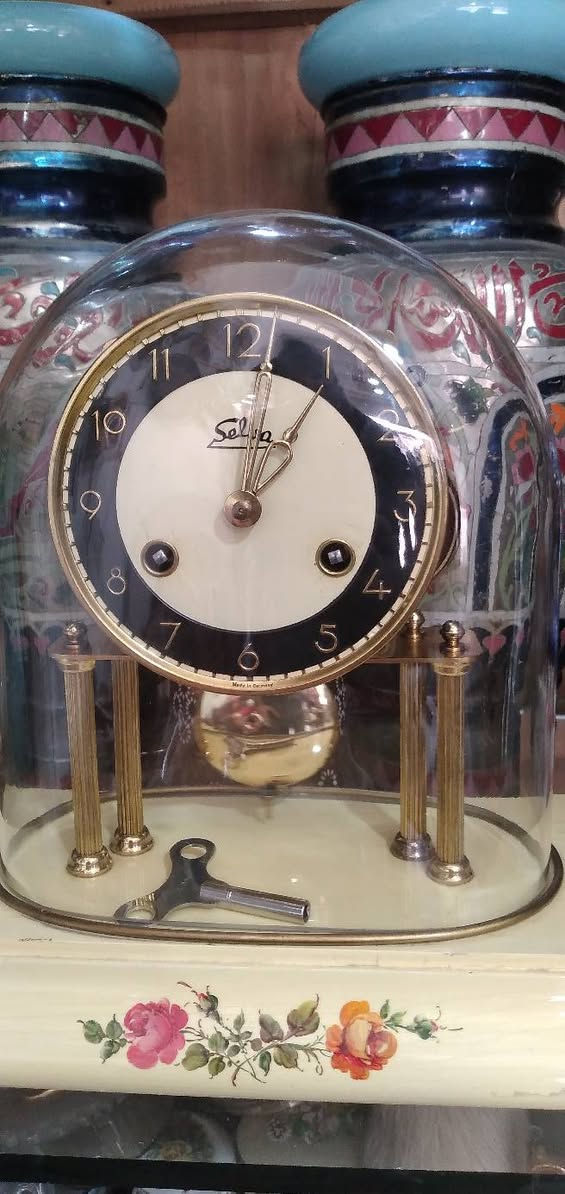 ساعة مكتبية أو رفّية (Mantel/Table Clock) كلاسيكية مميزة.  
​العلامة التجارية:   اسم الساعه "Selva"، وهي علامة تجارية ألمانية معروفة جداً في مجال قطع غيار الساعات، ومجموعات تركيب الساعات، والساعات الميكانيكية التقليدية بجودة عالية.
​المنشأ:  أسفل الرقم 6 عبارة صغيرة  مكتوب "Made in Germany" (صنع في ألمانيا)، وهو أمر متوافق تماماً مع العلامة التجارية Selva.
​ التصميم والهيكل
​القبة الزجاجية (Glass Dome): الساعة مغطاة بقبة زجاجية شفافة منحنية (تُعرف بساعات القبة أو ساعات الذكرى السنوية أحياناً، وإن كانت هذه تعمل ببندول تقليدي وليس بندولاً دواراً). وظيفتها حماية الآلية الداخلية والميناء من الأتربة.
​الميناء (Dial): يتميز بتصميم ثنائي اللون (حلقة خارجية سوداء تحتوي على أرقام عربية مذهبة واضحة، ومركز دائري بلون كريمي/أبيض مطفأ).
​الأعمدة والقاعدة: ترتكز الساعة على أعمدة نحاسية مضلعة مذهبة فوق قاعدة بيضاء أو كريمية المزينة بنقوش وردية ناعمة مرسومة باليد، مما يعطيها طابعاً أنثوياً أو ريفياً كلاسيكياً (Shabby Chic / Vintage style).
​الآلية والتشغيل (الميكانيكا)
​المفاتيح وثقوب التعبئة: يحتوي الميناء على ثقبين للتعبئة (Winding holes) بجانب الرقمين 4 و 8. هذا يعني أن الساعة تحتوي على نابضين (زُنبركين): واحد لتشغيل حركة عقارب الساعة، والآخر مخصص لآلية الرنين (الدق أو الجرس).
​مفتاح التعبئة:  مفتاح التعبئة المعدني الأصلي مستلقياً على قاعدة الساعة داخل القبة الزجاجية.
​البندول (Pendulum): بندول نحاسي لامع أسفل الميناء، وهو المسؤول عن تنظيم الوقت عبر حركته الاهتزازية يميناً ويساراً.
​ الحالة العامة  الساعة في حالة جيدة جداً ونظيفة. النحاس يحتفظ بلمعانه، والزجاج سليم لا تظهر فيه شروخ واضحة، والرسومات على القاعدة بحالة ممتازة.
​القيمة: هذا النوع من الساعات يجمع بين القيمة الوظيفية والجمالية كقطع ديكور "أنتيك" أو عتيقة مرغوبة لمحبي الساعات الألمانية الميكانيكية.


**إذا كنت صاحب هذا الإعلان وتريد حذفه لأي سبب، رجاءا أرسل رسالة إلى الدعم الفني**