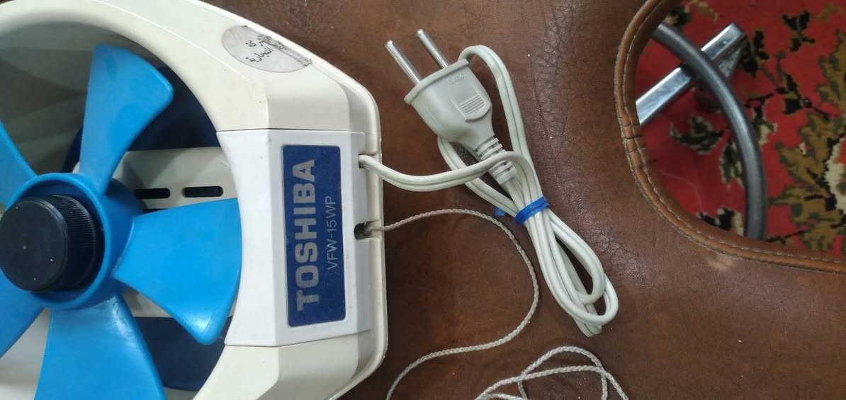 ***********
مروحة تهوية (شفاط) من نوع توشيبا (Toshiba)، وتتميز بتصميمها الكلاسيكي المتين الذي يركز على الكفاءة التشغيلية المباشرة.
​العلامة التجارية: توشيبا (Toshiba)
​الموديل: VFW-15WP.
​التصميم: مروحة شفط جدارية صغيرة الحجم، عادة ما تُستخدم في المطابخ أو الحمامات لتجديد الهواء.
​المكونات: 
​الشفرات: تحتوي على 5 ريش بلاستيكية باللون الأزرق، مصممة بتناظر لتوفير تدفق هواء هادئ ومنتظم.
​الهيكل: إطار بلاستيكي بلون كريمي/أبيض مائل للصفرة، وهو نمط شائع في الأجهزة اليابانية الكلاسيكية.
​نظام التشغيل: سلك سحب (Pull Cord) للتشغيل والإيقاف، وهو نظام ميكانيكي بسيط وعملي.
​التوصيل: سلك كهربائي ثنائي الرأس (Plug).
​الاستخدام والتركيب:
​هذا الموديل مخصص للتركيب داخل فتحات دائرية في الجدران أو النوافذ.
​يُعرف هذا النوع من إنتاج توشيبا بالاعتمادية العالية وطول العمر الافتراضي للمحرك، وهو ما يتماشى مع الاهتمام بالصناعات والمعدات ذات الطابع الفني والهندسي المتين.
​ بلد المنشأ اليابان ،
