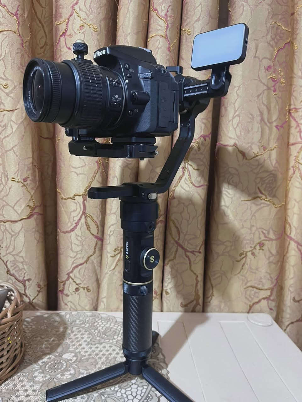 مكلف بل نشر
1. الكاميرا والعدسة (Body & Lens)
 نوع الكاميرا: Nikon D5300 (كاميرا احترافية من فئة DSLR معروفة جداً بموثوقيتها).
 دقة وميزات الكاميرا:
 مستشعر بدقة 24.2 ميجابكسل يعطي صوراً عالية الدقة وتفاصيل حادة.
 شاشة خلفية متحركة (تفتح وتفتر) وهي ممتازة جداً لتصوير الفيديو والـ Vlogs أو السيلفي.
 دعم تصوير فيديو بدقة Full HD (1080p) بسرعة تصل إلى 60 إطار في الثانية، مما يعطي سلاومة ونعومة بالفيديو.
 تحتوي على Wi-Fi مدمج لنقل الصور مباشرة للموبايل.
 العدسة (Lens): عدسة نيكون الأساسية AF-S NIKKOR 18-55mm f/3.5-5.6G VR.
 ميزة الزووم: مثل ما ذكرتِ، الصورة تتكبر وتتصغر (من 18mm للقطات الواسعة إلى 55mm للقطات القريبة والبورتريه).
 مانع اهتزاز (VR): العدسة مجهزة بنظام تقليل الاهتزاز الاهتزاز البصري لضمان صور واضحة بدون غواش حتى أثناء الحركة.
2. مانع الاهتزاز الاحترافي (Gimbal)
 النوع الموديل: Zhiyun Crane 2S (واضح من الشعار المكتوب باللون الذهبي والكتابة على يد المانع).
 وظيفة الجهاز: هذا الجهاز مخصص للمصورين وصناع المحتوى المحترفين؛ حيث يوازن الكاميرا تماماً ويمنع أي اهتزاز أثناء المشي أو الركض، ليظهر الفيديو ناعماً وسلساً مثل تصوير الأفلام السينمائية.
 المميزات الظاهرة بالصور:
 يد كربون فايبر (Carbon Fiber) خفيفة وقوية.
 أقفال أمان للمحاور (Axis Locks) تسهل عملية الموازنة والتنقل (كما في ⁠image.png⁠ و ⁠image_6.png⁠).
 قاعدة ترايبود (Tripod) سفلية صغيرة لثبات المانع على الأسطح.
3. الملحقات الإضافية (Accessories)
 إضاءة LED مدمجة: مثبتة أعلى مانع الاهتزاز (تظهر بوضوح في ⁠image_2.png⁠ و ⁠image_4.png⁠)؛ توفر إضاءة مستمرة وممتازة لتصوير الوجوه أو الفيديوهات في الأماكن المظلمة.
 حقيبة الحمل (Case): تظهر جزئياً في الخلفية بـ ⁠image_10.png⁠ وهي الحقيبة الأصلية لمانع الاهتزاز لحمايته أثناء النقل ***********
