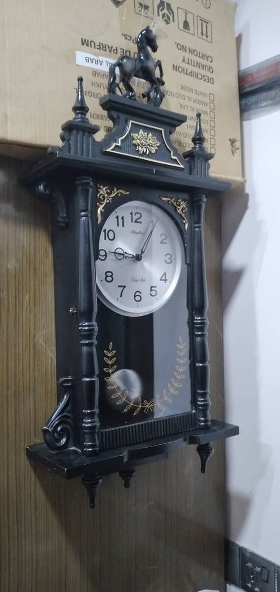 ساعة حائط بـ بندول (رقاص) من إنتاج شركة Rhythm اليابانية، وهي شركة عريقة معروفة بدقة محركاتها وتصاميمها الكلاسيكية.
​
​ هيكل الخارجي
​طراز: كلاسيكي مستوحى من طراز "منظم فيينا" (Vienna Regulator)، والذي يتميز بشكل طولي مع واجهة زجاجية تسمح برؤية البندول.
​اللون: الهيكل أسود المطفي مع لمسات تزيينية باللون الذهبي.
​ يوجد تمثال لحصان واثق، وهو عنصر تزييني تقليدي يمنح الساعة مظهراً فخماً.
​الأعمدة: على جانبي الساعة توجد أعمدة محفورة تعطي طابعاً معمارياً قديماً.
​النقوش: توجد زخارف نباتية (أزهار) في الجزء العلوي، وإكليل من الأوراق الذهبية على الزجاج السفلي يحيط بمجال حركة البندول.
​العلامة التجارية:  Rhythm   الموديل الذي يتميز بنغمات أو دقات معينة.
​ساعة بمحرك "كوارتز" (Quartz) دقيق.
​دق كل ساعة (أو نصف ساعة) بنغمات شهيرة مثل "Westminster


**إذا كنت صاحب هذا الإعلان وتريد حذفه لأي سبب، رجاءا أرسل رسالة إلى الدعم الفني**