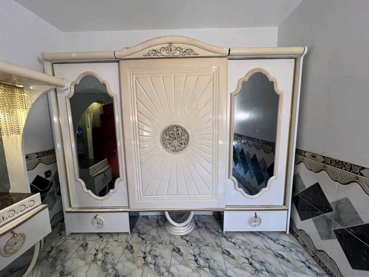 غرفة نوم تركية بتصميم فاخر وجميل، تتكون من سرير مزود بستارة شبكية وستائر مضاءة، بالإضافة إلى خزانة كبيرة مع مرايا. التصميم يجمع بين اللونين الأبيض والذهبي مع تغطية السرير بلون أحمر فاقع، مناسب لإضافة لمسة من الأناقة والرفاهية لغرفتك.