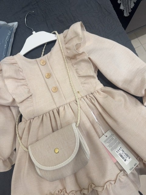 فستان بناتي بتصميم جميل مع حقيبة صغيرة مرفقة. لونه بيج ونسيجه ناعم، مثالي للمناسبات الخاصة. الأزرار الأمامية تضيف له لمسة أنيقة. متوفر بمجموعة مقاسات للأطفال.