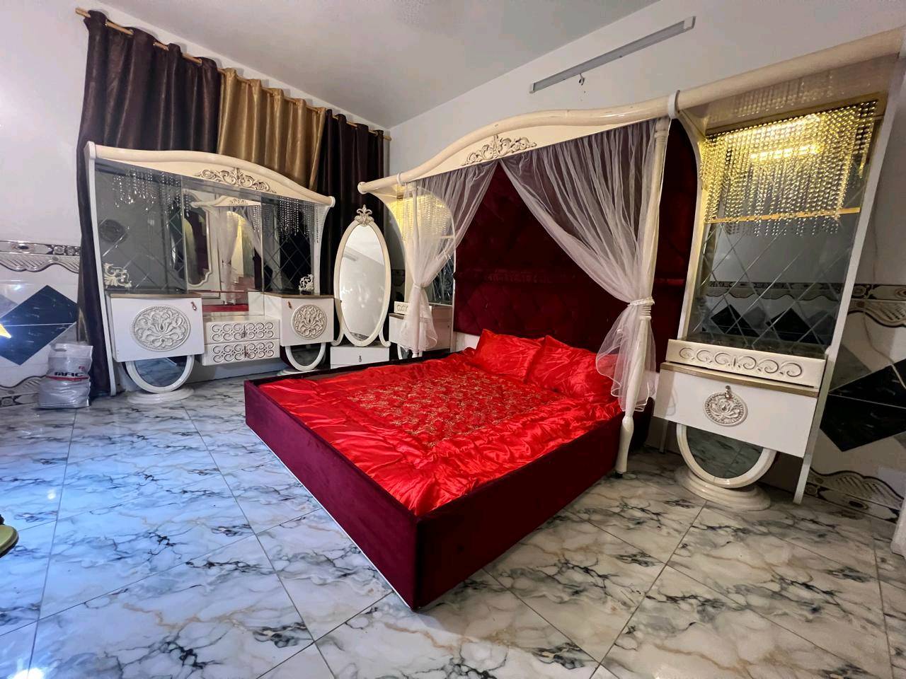 غرفة نوم تركية بتصميم فاخر وجميل، تتكون من سرير مزود بستارة شبكية وستائر مضاءة، بالإضافة إلى خزانة كبيرة مع مرايا. التصميم يجمع بين اللونين الأبيض والذهبي مع تغطية السرير بلون أحمر فاقع، مناسب لإضافة لمسة من الأناقة والرفاهية لغرفتك.