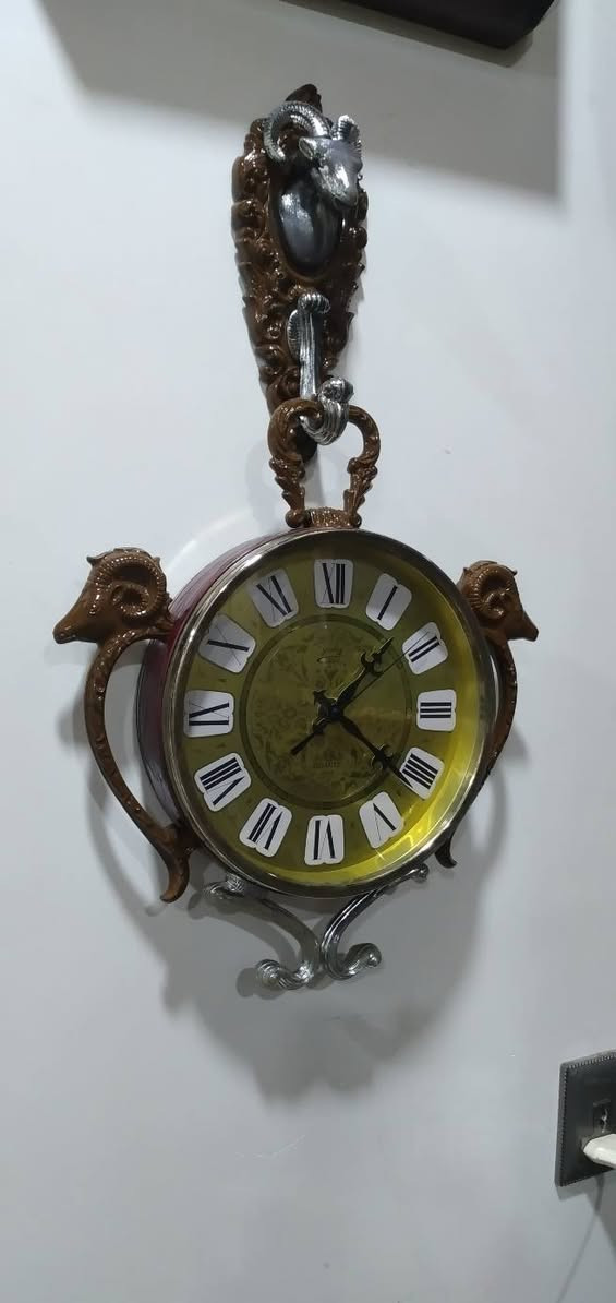 ساعة حائط مميزة جداً وتحمل طابعاً كلاسيكياً عتيقاً (Vintage/Retro)، وتحديداً من الساعات التي اشتهرت بها شركة "ريذم" (Rhythm) أو "كاسيو" (Casio) اليابانية في فترة السبعينيات والثمانينيات، والتي كانت تعتمد على تصاميم وزخارف بلاستيكية أو معدنية تحاكي الأنماط الأوروبية الكلاسيكية الكثيفة. مواصفات 
​ التصميم والهيكل الخارجي (The Aesthetic)
​ التصميم مستوحى من شكل "ساعة الجيب الكلاسيكية العتيقة" من حيث وجود الحلقة العلوية الكبيرة والمقبض، ولكن تمت إضافة عناصر زخرفية بارزة تمنحها طابعاً ملكياً أو ريفياً أوروبياً (Baroque / Rococo Style).
​التماثيل والزخارف: * رأس الكبش (أو الوعل): نلاحظ وجود رأس كبش بقرون دائرية مميزة في أعلى الساعة باللون الفضي المحاط بإطار بني مزخرف، وتتكرر رؤوس الكباش على الجانبين الأيمن والأيسر كحوامل أو مقابض جانبية. هذا الرمز غالباً ما يرمز للقوة أو يرتبط بتصاميم الساعات الجدارية المخصصة لغرف الضيوف أو المكاتب الفخمة.
​القاعدة السفلية: مزينة بزخرفة معدنية ملتوية باللون الفضي (Scrollwork) لتعطي توازناً بصرياً مع الجزء العلوي.
 الميناء والعقارب (The Dial & Hands)
​الخلفية (الميناء): تأتي باللون الأخضر الزيتوني المتدرج إلى الأصفر (Sunburst/Gradient Effect)، وهو لون كلاسيكي شهير جداً في حقبة السبعينيات. يوجد في منتصف الميناء نقوش وزخارف نباتية ناعمة ملتفة (Filigree Pattern).
​الأرقام: أرقام رومانية كبيرة باللون الأسود موضوعة داخل مربعات أو مخروطيات بيضاء بارزة، مما يسهل قراءتها ويمنح الميناء بُعداً ثلاثي الأبعاد.
​العقارب: العقارب مصممة على الطراز الكلاسيكي (Retro/Ornate Hands) مع وجود عقرب ثوانٍ نحيف.
​الكتابة: يظهر في النصف السفلي من الميناء كلمة "QUARTZ"، مما يؤكد أن الساعة تعمل بحركة الكوارتز الحديثة (التي تعتمد على البطارية) وليست حركة ميكانيكية بالزنبرك، وهو ما كان ثورة في دقة الوقت خلال تلك الحقبة.
 التحليل الفني والمنشأ
​الحركة الداخلية: تعمل الساعة بنظام الكوارتز (Battery-powered Quartz Movement)، وغالباً ما تكون الماكينة الداخلية يابانية الصنع (مثل ماكينات Rhythm الموثوقة جداً في امتصاص الطاقة وتعمير البطارية).
​المواد المستخدمة: الهيكل الخارجي مصنع من البلاستيك المقوى عالي الجودة (Hard Injection-Molded Plastic) الذي كان يُدهن بطبقات تحاكي الخشب الداكن أو النحاس والمعدن الفضي، وهي تقنية برعت فيها المصانع اليابانية لإنتاج ساعات حائط خفيفة الوزن ذات مظهر فخم وثقيل.


**إذا كنت صاحب هذا الإعلان وتريد حذفه لأي سبب، رجاءا أرسل رسالة إلى الدعم الفني**