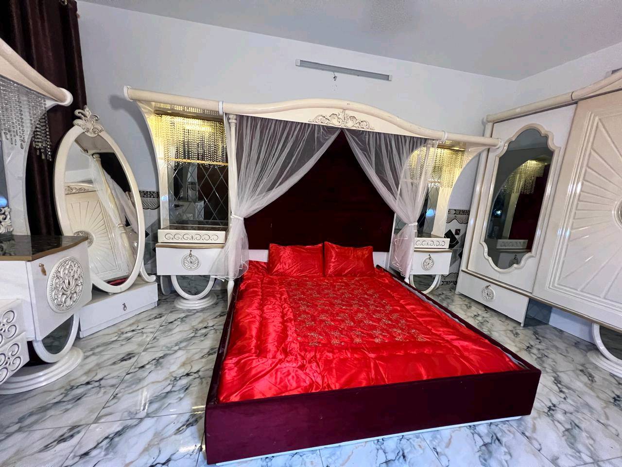 غرفة نوم تركية بتصميم فاخر وجميل، تتكون من سرير مزود بستارة شبكية وستائر مضاءة، بالإضافة إلى خزانة كبيرة مع مرايا. التصميم يجمع بين اللونين الأبيض والذهبي مع تغطية السرير بلون أحمر فاقع، مناسب لإضافة لمسة من الأناقة والرفاهية لغرفتك.