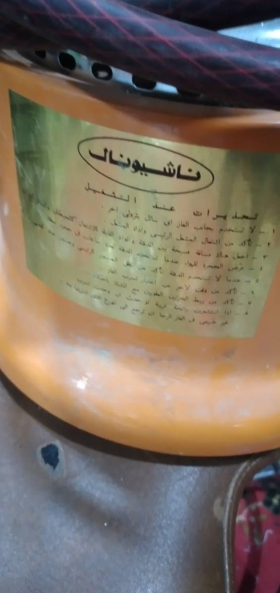 موقد غازي محمول (أو دفاية غازية متعددة الاستخدامات) من إنتاج شركة National (ناشيونال) اليابانية العريقة، ويعود تصميمه إلى فترة السبعينات أو الثمانينات.
​يتميز هذا التصميم بالجمع بين وظيفتين: التدفئة بفضل المشبك الشبكي الحراري، والطبخ/التسخين بفضل الحامل العلوي المتين.
​إليك تحليلاً تفصيلياً للمكونات والتصميم الهندسي لهذا الجهاز الكلاسيكي:

​١. الهيكل الخارجي والتصميم العام

​اللون والقاعدة: يأتي الهيكل السفلي باللون البرتقالي الكلاسيكي المميز للأجهزة المنزلية في تلك الحقبة. القاعدة دائرية وعريضة تضمن ثبات الجهاز على الأرض أو الطاولة لمنع الانقلاب.

​لوحة التحكم والسويتش: في المقدمة توجد لوحة تحكم سوداء تحمل شعار National باللون الأحمر والأبيض التقليدي. يحتوي على مفتاح دوار رمادي كبير للتحكم في تدفق الغاز (التشغيل والإطفاء "OFF" وتعديل مستوى اللهب).

​٢. منظومة الاحتراق والتدفئة (الجزء العلوي)

​غرفة الاحتراق الشبكية: يوجد في المنتصف أسطوانة شبكية معدنية دقيقة. عندما يشتعل الغاز، تسخن هذه الشبكة حتى التوهج (اللون الأحمر)، مما يساعد على تحويل اللهب الأزرق المباشر إلى حرارة إشعاعية لتوزيع التدفئة بشكل أفضل في المكان.

​شبك الحماية (المشبك السلكي): يحيط بغرفة الاحتراق قفص سلكي معدني سميك لحماية المستخدم من الملامسة المباشرة للأسطح شديدة السخونة، وهو مصمم بشكل عمودي متين.

​حامل الأواني العلوي (المنصب): في القمة يوجد حامل زهر (أو معدن ثقيل) بأربعة أطراف مضلعة، مصمم لتحمل الأواني، الأباريق، أو دلال القهوة والشاي، مما يجعله موقداً عملياً جداً. نظام التغذية والأمان

​خرطوم الغاز:  خرطوم غاز سميك مقوى ومحاط بملف زنبركي لحمايته من الانثناء الحاد أو التلف عند مخرج الموقد. هذا النوع يعتمد على التغذية من أسطوانة غاز خارجية عبر منظم (خافض ضغط).

​ملصق الإرشادات: يوجد على الجانب الأيسر ملصق أصفر يحتوي على التعليمات والتحذيرات الفنية الخاصة بالتشغيل والأمان (غالباً باللغة العربية أو الإنجليزية حسب السوق الموجه إليه).

​القيمة التراثية والعملية

​أجهزة ناشيونال اليابانية من هذه الحقبة معروفة بجودة التصنيع الفائقة ("Japan Made") والاعتمادية العالية؛ حيث تعيش موادها (مثل الصاج المطلي والمفاتيح الميكانيكية) لعقود طويلة دون تلف إذا ما تم الحفاظ عليها وتنظيف قنوات الغاز فيها بانتظام.
​هل تود معرفة تفاصيل أكثر حول كيفية صيانة الفونية (مخرج الغاز) أو التأكد من سلامة صمامات الأمان فيه؟


**إذا كنت صاحب هذا الإعلان وتريد حذفه لأي سبب، رجاءا أرسل رسالة إلى الدعم الفني**