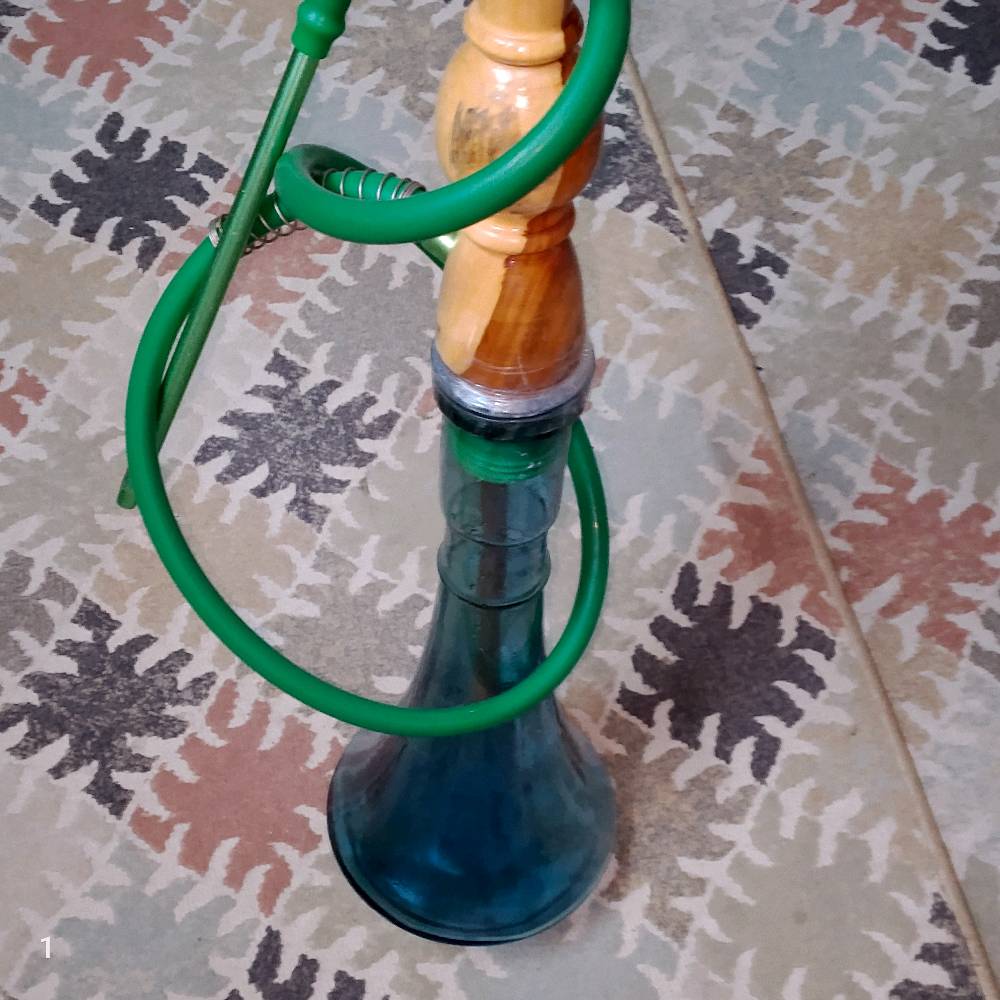 نركيلة خشبية ذات قاعدة زجاجية بلون أزرق وتظهر في حالة عامة جيدة. مزودة بخرطوم أخضر اللون وتتميز بتصميم جذاب ومتين. مناسبة للجلسات الخاصة وتضيف لمسة مميزة للأجواء.