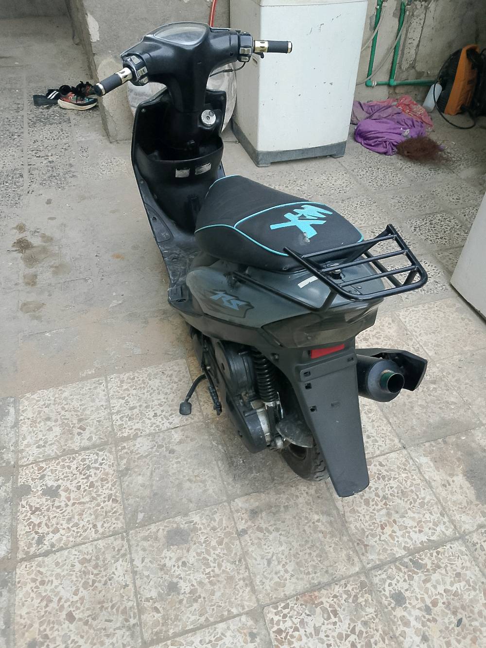 دراجة نارية نوع سكوتر بحالة جيدة، اللون رمادي مع شعار مميز على المقعد. الموديل مجهز بحاملة خلفية للأغراض، والمقابض بلون ذهبي تضيف لمسة أنيقة. مناسبة للتنقل اليومي وسهلة القيادة في الشوارع الضيقة.