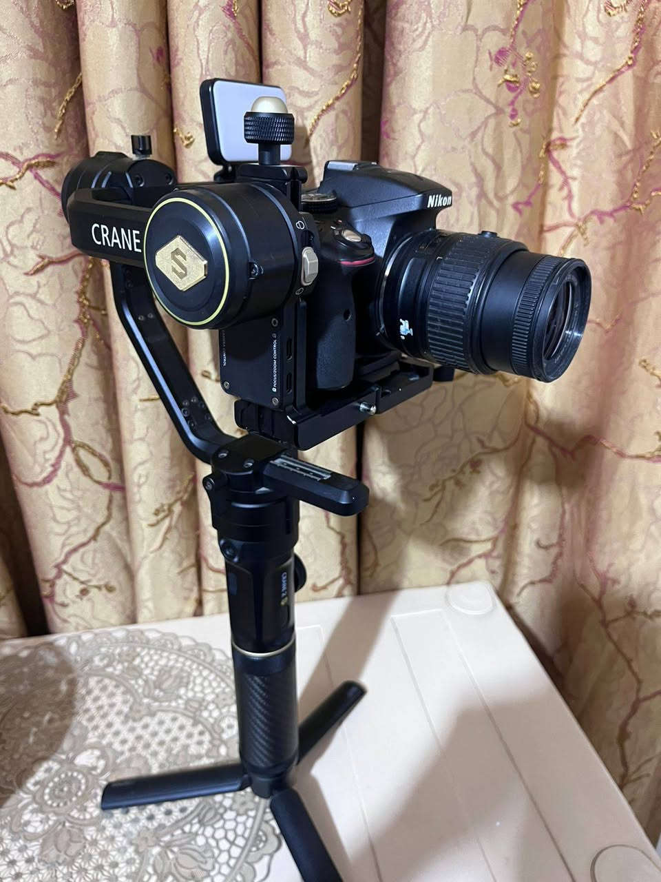 1. الكاميرا والعدسة (Body & Lens)
نوع الكاميرا: Nikon D5300 (كاميرا احترافية من فئة DSLR معروفة جداً بموثوقيتها).
 دقة وميزات الكاميرا:
 مستشعر بدقة 24.2 ميجابكسل يعطي صوراً عالية الدقة وتفاصيل حادة.
 شاشة خلفية متحركة (تفتح وتفتر) وهي ممتازة جداً لتصوير الفيديو والـ Vlogs أو السيلفي.
 دعم تصوير فيديو بدقة Full HD (1080p) بسرعة تصل إلى 60 إطار في الثانية، مما يعطي سلاومة ونعومة بالفيديو.
 تحتوي على Wi-Fi مدمج لنقل الصور مباشرة للموبايل.
 العدسة (Lens): عدسة نيكون الأساسية AF-S NIKKOR 18-55mm f/3.5-5.6G VR.
 ميزة الزووم: مثل ما ذكرتِ، الصورة تتكبر وتتصغر (من 18mm للقطات الواسعة إلى 55mm للقطات القريبة والبورتريه).
 مانع اهتزاز (VR): العدسة مجهزة بنظام تقليل الاهتزاز الاهتزاز البصري لضمان صور واضحة بدون غواش حتى أثناء الحركة.
2. مانع الاهتزاز الاحترافي (Gimbal)
 النوع الموديل: Zhiyun Crane 2S (واضح من الشعار المكتوب باللون الذهبي والكتابة على يد المانع).
 وظيفة الجهاز: هذا الجهاز مخصص للمصورين وصناع المحتوى المحترفين؛ حيث يوازن الكاميرا تماماً ويمنع أي اهتزاز أثناء المشي أو الركض، ليظهر الفيديو ناعماً وسلساً مثل تصوير الأفلام السينمائية.
 المميزات الظاهرة بالصور:
 يد كربون فايبر (Carbon Fiber) خفيفة وقوية.
 أقفال أمان للمحاور (Axis Locks) تسهل عملية الموازنة والتنقل (كما في ⁠image.png⁠ و ⁠image_6.png⁠).
 قاعدة ترايبود (Tripod) سفلية صغيرة لثبات المانع على الأسطح.
3. الملحقات الإضافية (Accessories)
 إضاءة LED مدمجة: مثبتة أعلى مانع الاهتزاز (تظهر بوضوح في ⁠image_2.png⁠ و ⁠image_4.png⁠)؛ توفر إضاءة مستمرة وممتازة لتصوير الوجوه أو الفيديوهات في الأماكن المظلمة.
 حقيبة الحمل (Case): تظهر جزئياً في الخلفية بـ ⁠image_10.png⁠ وهي الحقيبة الأصلية لمانع الاهتزاز لحمايته أثناء النقل.


**إذا كنت صاحب هذا الإعلان وتريد حذفه لأي سبب، رجاءا أرسل رسالة إلى الدعم الفني**