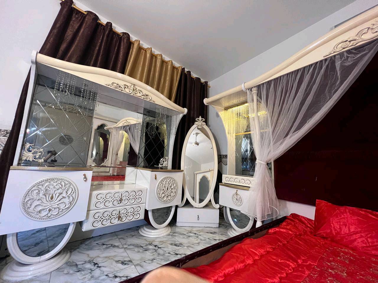 غرفة نوم تركية بتصميم فاخر وجميل، تتكون من سرير مزود بستارة شبكية وستائر مضاءة، بالإضافة إلى خزانة كبيرة مع مرايا. التصميم يجمع بين اللونين الأبيض والذهبي مع تغطية السرير بلون أحمر فاقع، مناسب لإضافة لمسة من الأناقة والرفاهية لغرفتك.