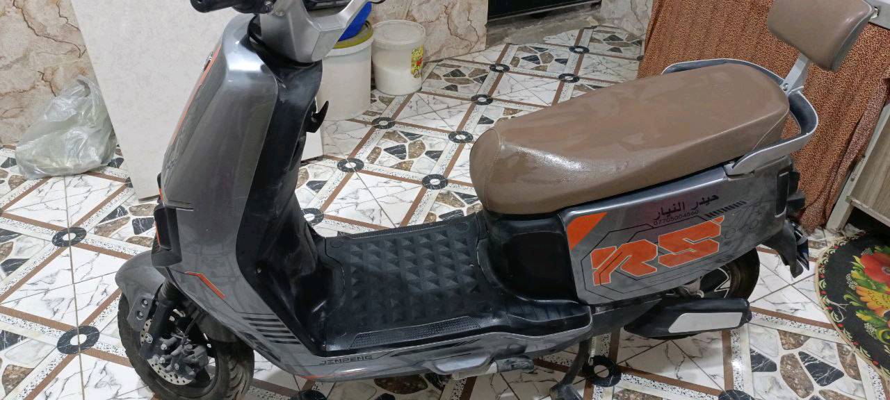 دراجة نارية كهربائية حديثة، بحالة جيدة جداً، لونها رمادي مع تفاصيل برتقالية مميزة. تحتوي على بطارية قوية لتوفير تجربة قيادة سلسة واقتصادية. مناسبة للتنقل اليومي داخل المدينة.