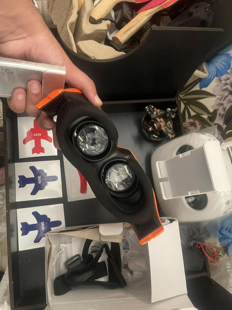 1. نظارة الطيران الافتراضي (FPV Goggles)
 الوصف: هي الأداة التي تحملها بيدك باللونين الأسود والبرتقالي.
 الوظيفة: تحتوي على شاشة وعدسات داخلية تعرض لك بثاً مباشراً وحياً من الكاميرا المثبتة على مقدمة الدرون، لتعيش تجربة الطيران وكأنك داخل مقصورة القيادة.
 الملحقات: يظهر أسفلها الحزام المطاطي الأسود (Headstrap) الخاص بتثبيت النظارة على الرأس داخل علبتها الكرتونية.


**إذا كنت صاحب هذا الإعلان وتريد حذفه لأي سبب، رجاءا أرسل رسالة إلى الدعم الفني**