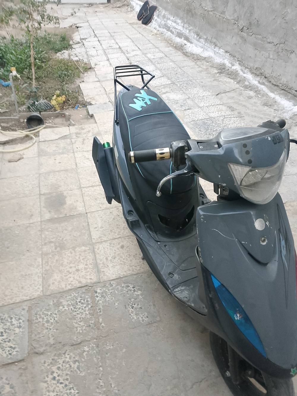 دراجة نارية نوع سكوتر بحالة جيدة، اللون رمادي مع شعار مميز على المقعد. الموديل مجهز بحاملة خلفية للأغراض، والمقابض بلون ذهبي تضيف لمسة أنيقة. مناسبة للتنقل اليومي وسهلة القيادة في الشوارع الضيقة.