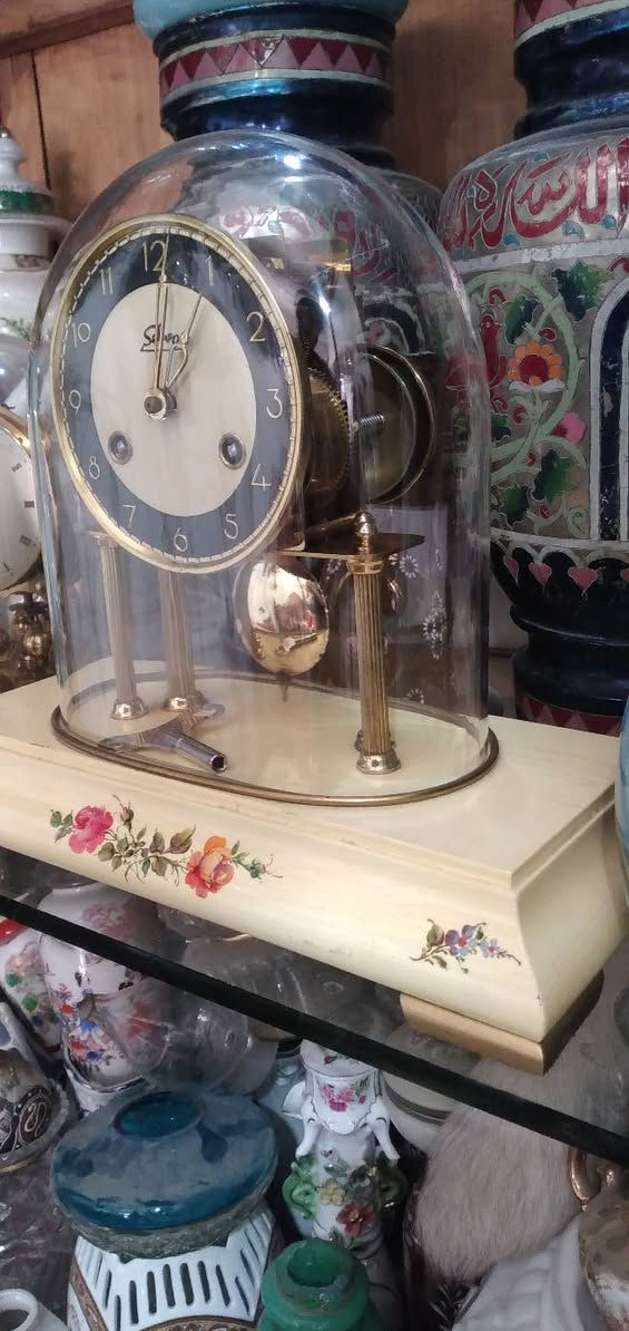 ساعة مكتبية أو رفّية (Mantel/Table Clock) كلاسيكية مميزة.  
​العلامة التجارية:   اسم الساعه "Selva"، وهي علامة تجارية ألمانية معروفة جداً في مجال قطع غيار الساعات، ومجموعات تركيب الساعات، والساعات الميكانيكية التقليدية بجودة عالية.
​المنشأ:  أسفل الرقم 6 عبارة صغيرة  مكتوب "Made in Germany" (صنع في ألمانيا)، وهو أمر متوافق تماماً مع العلامة التجارية Selva.
​ التصميم والهيكل
​القبة الزجاجية (Glass Dome): الساعة مغطاة بقبة زجاجية شفافة منحنية (تُعرف بساعات القبة أو ساعات الذكرى السنوية أحياناً، وإن كانت هذه تعمل ببندول تقليدي وليس بندولاً دواراً). وظيفتها حماية الآلية الداخلية والميناء من الأتربة.
​الميناء (Dial): يتميز بتصميم ثنائي اللون (حلقة خارجية سوداء تحتوي على أرقام عربية مذهبة واضحة، ومركز دائري بلون كريمي/أبيض مطفأ).
​الأعمدة والقاعدة: ترتكز الساعة على أعمدة نحاسية مضلعة مذهبة فوق قاعدة بيضاء أو كريمية المزينة بنقوش وردية ناعمة مرسومة باليد، مما يعطيها طابعاً أنثوياً أو ريفياً كلاسيكياً (Shabby Chic / Vintage style).
​الآلية والتشغيل (الميكانيكا)
​المفاتيح وثقوب التعبئة: يحتوي الميناء على ثقبين للتعبئة (Winding holes) بجانب الرقمين 4 و 8. هذا يعني أن الساعة تحتوي على نابضين (زُنبركين): واحد لتشغيل حركة عقارب الساعة، والآخر مخصص لآلية الرنين (الدق أو الجرس).
​مفتاح التعبئة:  مفتاح التعبئة المعدني الأصلي مستلقياً على قاعدة الساعة داخل القبة الزجاجية.
​البندول (Pendulum): بندول نحاسي لامع أسفل الميناء، وهو المسؤول عن تنظيم الوقت عبر حركته الاهتزازية يميناً ويساراً.
​ الحالة العامة  الساعة في حالة جيدة جداً ونظيفة. النحاس يحتفظ بلمعانه، والزجاج سليم لا تظهر فيه شروخ واضحة، والرسومات على القاعدة بحالة ممتازة.
​القيمة: هذا النوع من الساعات يجمع بين القيمة الوظيفية والجمالية كقطع ديكور "أنتيك" أو عتيقة مرغوبة لمحبي الساعات الألمانية الميكانيكية.


**إذا كنت صاحب هذا الإعلان وتريد حذفه لأي سبب، رجاءا أرسل رسالة إلى الدعم الفني**