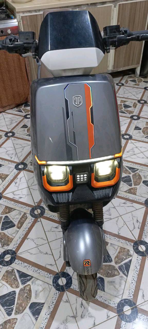 دراجة نارية كهربائية حديثة، بحالة جيدة جداً، لونها رمادي مع تفاصيل برتقالية مميزة. تحتوي على بطارية قوية لتوفير تجربة قيادة سلسة واقتصادية. مناسبة للتنقل اليومي داخل المدينة.
