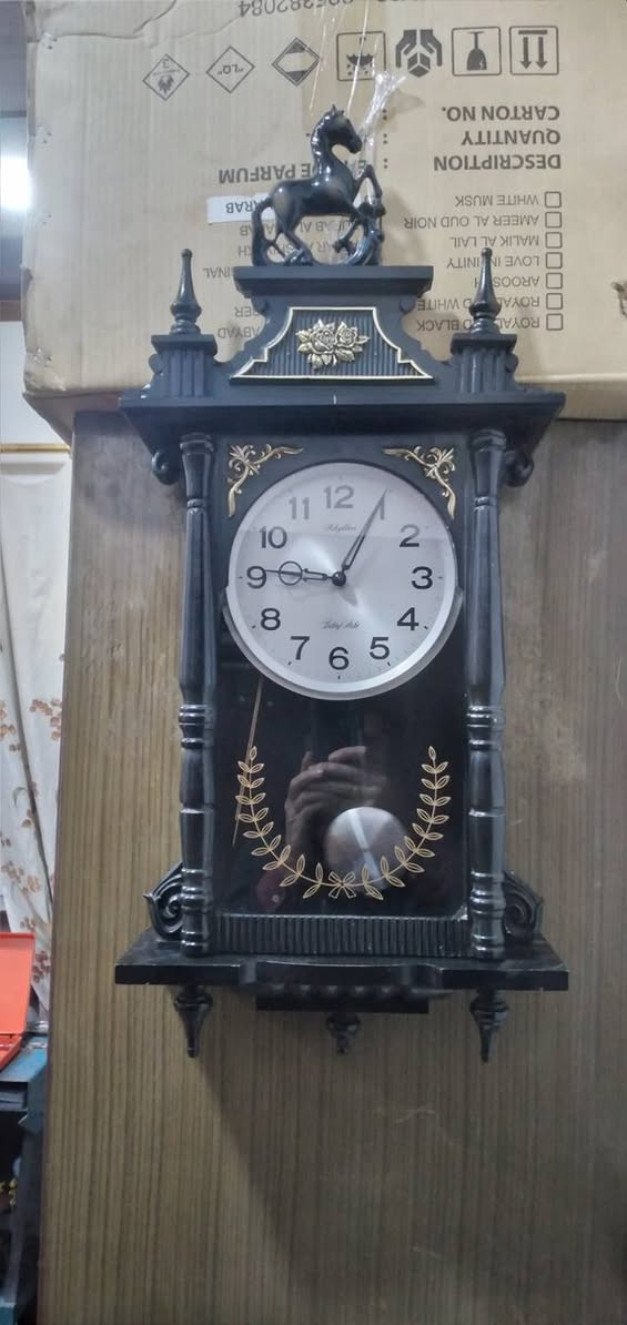 ساعة حائط بـ بندول (رقاص) من إنتاج شركة Rhythm اليابانية، وهي شركة عريقة معروفة بدقة محركاتها وتصاميمها الكلاسيكية.
​
​ هيكل الخارجي
​طراز: كلاسيكي مستوحى من طراز "منظم فيينا" (Vienna Regulator)، والذي يتميز بشكل طولي مع واجهة زجاجية تسمح برؤية البندول.
​اللون: الهيكل أسود المطفي مع لمسات تزيينية باللون الذهبي.
​ يوجد تمثال لحصان واثق، وهو عنصر تزييني تقليدي يمنح الساعة مظهراً فخماً.
​الأعمدة: على جانبي الساعة توجد أعمدة محفورة تعطي طابعاً معمارياً قديماً.
​النقوش: توجد زخارف نباتية (أزهار) في الجزء العلوي، وإكليل من الأوراق الذهبية على الزجاج السفلي يحيط بمجال حركة البندول.
​العلامة التجارية:  Rhythm   الموديل الذي يتميز بنغمات أو دقات معينة.
​ساعة بمحرك "كوارتز" (Quartz) دقيق.
​دق كل ساعة (أو نصف ساعة) بنغمات شهيرة مثل "Westminster


**إذا كنت صاحب هذا الإعلان وتريد حذفه لأي سبب، رجاءا أرسل رسالة إلى الدعم الفني**