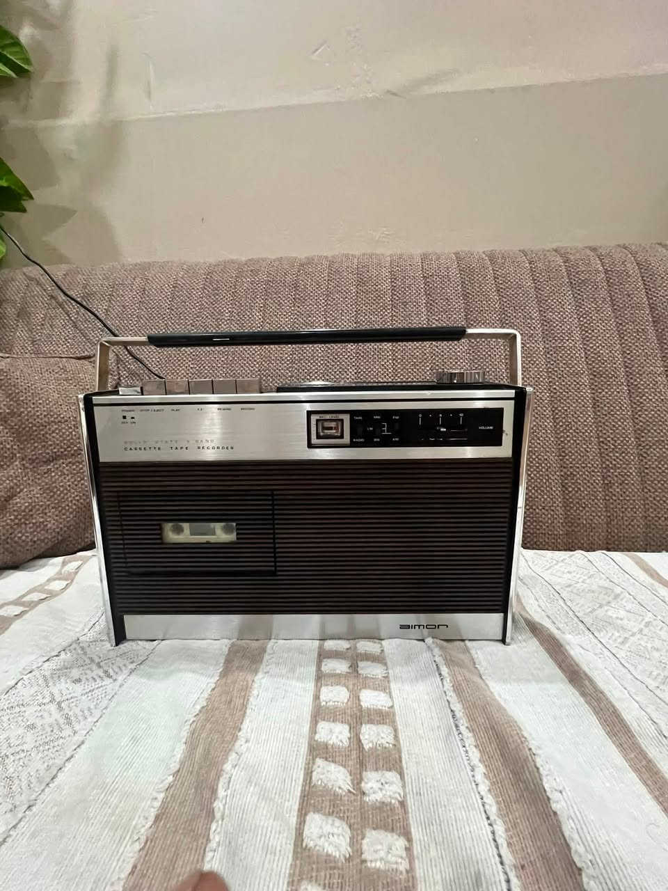 هذا جهاز راديو ومسجل كاسيت كلاسيكي مميز جداً يحمل العلامة التجارية Bimon.
إليك نظرة سريعة على تفاصيل هذا الجهاز الظاهرة في الصورة:
 المواصفات الأساسية: هو مسجل كاسيت وراديو ترانزستور عالي الثبات (Solid State) يدعم 4 موجات راديو مختلفة (4 Band) تشمل عادةً (FM, MW, LW, SW) لالتقاط الإشارات المحلية والقصيرة.
 التصميم: يتميز بالهيكل الكلاسيكي الأنيق الذي يدمج بين اللمسات المعدنية الفضية في الأعلى والأسفل، والواجهة المخططة ذات المظهر الخشبي الداكن (Woodgrain)، وهو أسلوب التصميم الذي كان سائداً ومحبوباً جداً في فترة السبعينيات وأوائل الثمانينيات.
 عناصر التحكم: يحتوي في الجزء العلوي على المفاتيح الميكانيكية التقليدية (Piano Keys) للتحكم بالمسجل (تشغيل، تقديم، إرجاع، إيقاف/إخراج)، بالإضافة إلى مؤشر صغير للإشارة وضبط الصوت والموجات على الجانب الأيمن.
هل للبيع نظيف جدا  ***********
