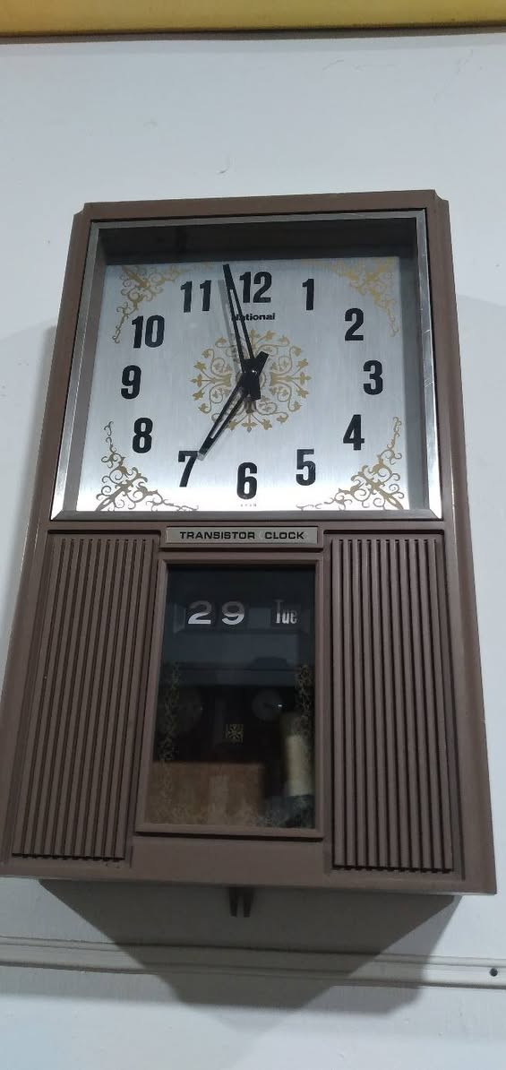 ***********
كلاسيكية رائعة جداً!  ساعة حائط يابانية عتيقة (Vintage) من إنتاج شركة National (ناشيونال - المعروفة حالياً باسم باناسونيك Panasonic)، وتحديداً من فئة ساعات "Transistor Clock" (ساعة ترانزستور) التي كانت شائعة جداً في فترة أواخر الستينيات والسبعينيات من القرن الماضي.
​الهيكل والتصميم الخارجي
​النمط المستوحى من الخمسينيات والستينيات (Mid-Century Modern): تتميز الساعة بهيكل مستطيل رأسي، مطلي باللون البني الكلاسيكي مع وجود خطوط رأسية (مشرشرة) على الجانبين السفليين تشبه فتحات المكبرات الصوتية في الراديو القديم، وهو أسلوب تصميمي كان دارجاً جداً ليعطي انطباعاً بالفخامة والدفء المنزلي.
​مينا الساعة (Dial): المينا يأتي بخلفية معدنية فضية لامعة (Brushed aluminum)، محاطة بزخارف ذهبية كلاسيكية في الأركان وفي المنتصف. الأرقام العربية كبيرة، سوداء، وواضحة جداً بخط سميك يسهل قراءته من مسافة بعيدة.
​العلامة التجارية والمنشأ:   شعار National أسفل الرقم 12، وفي الأسفل تماماً تحت الرقم 6 كُتبت عبارة JAPAN لتؤكد جودة الصناعة اليابانية في تلك الحقبة.
​ آلية العمل (الحركة والتكنولوجيا)
​Transistor Clock (ساعة الترانزستور): اللوحة الفضية الصغيرة أسفل المينا تحمل عبارة "TRANSISTOR CLOCK". هذه التكنولوجيا كانت طفرة في وقتها؛ فهي تمثل مرحلة انتقالية بين الساعات الميكانيكية بالكامل (التي تعتمد على الزنبرك والتعبئة اليدوية) وساعات الكوارتز الحديثة. تعمل هذه الساعة بالبطارية، حيث يقوم ترانزستور صغير بتنظيم النبضات الكهربائية لتحريك بندول مغناطيسي أو عجلة توازن، مما يحافظ على دقة الوقت دون الحاجة لتعبئتها يدوياً.
​البندول (Pendulum): في النافذة السفلية المحاطة بالإطار البني، يمكن رؤية جزء من البندول الداخلي الذي يتحرك يميناً ويساراً، وهو مغطى بزجاج شفاف يتيح رؤية الحركة الميكانيكية التي تعطي الساعة روحاً وجمالية خاصة.
​ الميزات الإضافية (التقويم)
​نافذة التاريخ واليوم (Day/Date Calendar): بجانب البندول في القسم السفلي، تحتوي الساعة على تقويم ميكانيكي يظهر اليوم والشهر (يظهر في الصورة رقم 29 واليوم "Tue" أي الثلاثاء). هذا النوع من التقويم يتغير تلقائياً مع حركة عقارب الساعة عند منتصف الليل.
​باختصار، هذه الساعة ليست مجرد أداة لمعرفة الوقت، بل هي قطعة فنية تجسد ذروة الصناعة الهندسية والجمالية اليابانية في السبعينيات، وتعتبر اليوم من القطع المميزة جداً لهواة جمع المقتنيات والأنتيكات الإلكترو-ميكانيكية.
