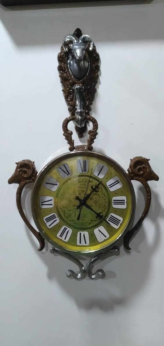 ساعة حائط مميزة جداً وتحمل طابعاً كلاسيكياً عتيقاً (Vintage/Retro)، وتحديداً من الساعات التي اشتهرت بها شركة "ريذم" (Rhythm) أو "كاسيو" (Casio) اليابانية في فترة السبعينيات والثمانينيات، والتي كانت تعتمد على تصاميم وزخارف بلاستيكية أو معدنية تحاكي الأنماط الأوروبية الكلاسيكية الكثيفة. مواصفات 
​ التصميم والهيكل الخارجي (The Aesthetic)
​ التصميم مستوحى من شكل "ساعة الجيب الكلاسيكية العتيقة" من حيث وجود الحلقة العلوية الكبيرة والمقبض، ولكن تمت إضافة عناصر زخرفية بارزة تمنحها طابعاً ملكياً أو ريفياً أوروبياً (Baroque / Rococo Style).
​التماثيل والزخارف: * رأس الكبش (أو الوعل): نلاحظ وجود رأس كبش بقرون دائرية مميزة في أعلى الساعة باللون الفضي المحاط بإطار بني مزخرف، وتتكرر رؤوس الكباش على الجانبين الأيمن والأيسر كحوامل أو مقابض جانبية. هذا الرمز غالباً ما يرمز للقوة أو يرتبط بتصاميم الساعات الجدارية المخصصة لغرف الضيوف أو المكاتب الفخمة.
​القاعدة السفلية: مزينة بزخرفة معدنية ملتوية باللون الفضي (Scrollwork) لتعطي توازناً بصرياً مع الجزء العلوي.
 الميناء والعقارب (The Dial & Hands)
​الخلفية (الميناء): تأتي باللون الأخضر الزيتوني المتدرج إلى الأصفر (Sunburst/Gradient Effect)، وهو لون كلاسيكي شهير جداً في حقبة السبعينيات. يوجد في منتصف الميناء نقوش وزخارف نباتية ناعمة ملتفة (Filigree Pattern).
​الأرقام: أرقام رومانية كبيرة باللون الأسود موضوعة داخل مربعات أو مخروطيات بيضاء بارزة، مما يسهل قراءتها ويمنح الميناء بُعداً ثلاثي الأبعاد.
​العقارب: العقارب مصممة على الطراز الكلاسيكي (Retro/Ornate Hands) مع وجود عقرب ثوانٍ نحيف.
​الكتابة: يظهر في النصف السفلي من الميناء كلمة "QUARTZ"، مما يؤكد أن الساعة تعمل بحركة الكوارتز الحديثة (التي تعتمد على البطارية) وليست حركة ميكانيكية بالزنبرك، وهو ما كان ثورة في دقة الوقت خلال تلك الحقبة.
 التحليل الفني والمنشأ
​الحركة الداخلية: تعمل الساعة بنظام الكوارتز (Battery-powered Quartz Movement)، وغالباً ما تكون الماكينة الداخلية يابانية الصنع (مثل ماكينات Rhythm الموثوقة جداً في امتصاص الطاقة وتعمير البطارية).
​المواد المستخدمة: الهيكل الخارجي مصنع من البلاستيك المقوى عالي الجودة (Hard Injection-Molded Plastic) الذي كان يُدهن بطبقات تحاكي الخشب الداكن أو النحاس والمعدن الفضي، وهي تقنية برعت فيها المصانع اليابانية لإنتاج ساعات حائط خفيفة الوزن ذات مظهر فخم وثقيل.


**إذا كنت صاحب هذا الإعلان وتريد حذفه لأي سبب، رجاءا أرسل رسالة إلى الدعم الفني**