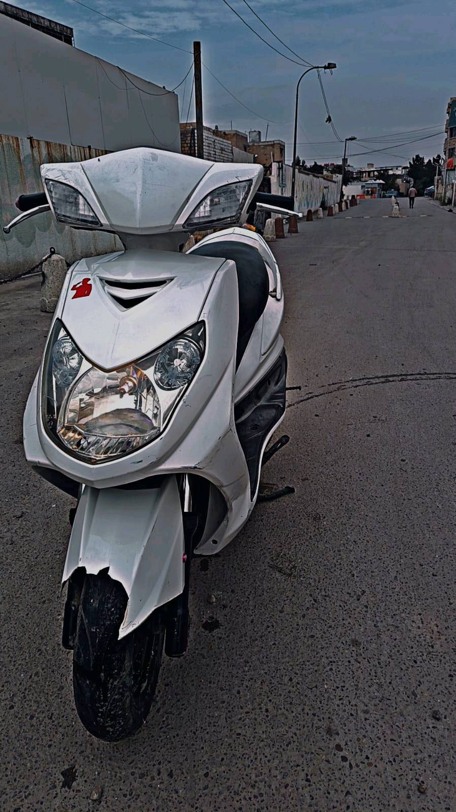 دراجة نارية نوع سكوتر لون أبيض بحالة جيدة، مثالية للتنقل داخل المدينة. تصميمها أنيق ومزودة بأضواء أمامية ساطعة لسهولة القيادة في الليل. مناسبة للي يبحث عن وسيلة نقل اقتصادية وعملية.