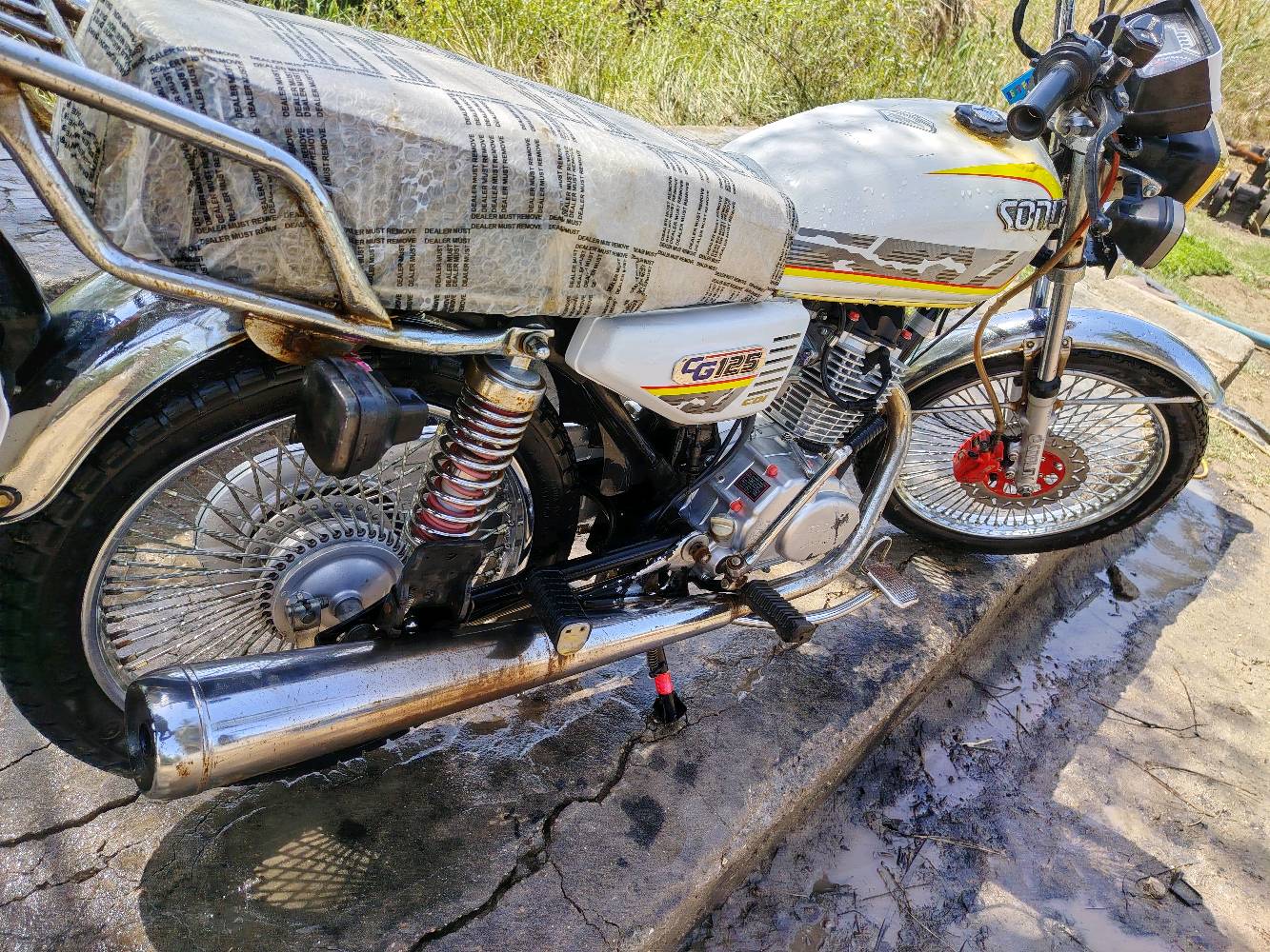 دراجة نارية نوع دايون CG125 بحالة ممتازة، لونها أبيض مع رسومات جانبية. الدراجة نظيفة ومجهزة بالكامل، مما يجعلها مثالية للاستخدام اليومي أو المشاوير الطويلة.