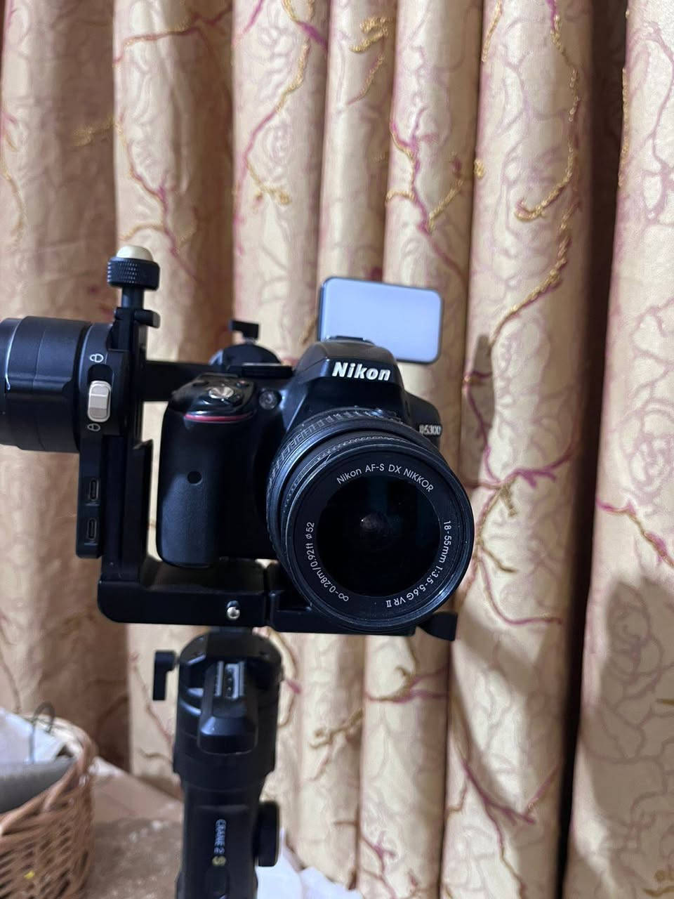 1. الكاميرا والعدسة (Body & Lens)
نوع الكاميرا: Nikon D5300 (كاميرا احترافية من فئة DSLR معروفة جداً بموثوقيتها).
 دقة وميزات الكاميرا:
 مستشعر بدقة 24.2 ميجابكسل يعطي صوراً عالية الدقة وتفاصيل حادة.
 شاشة خلفية متحركة (تفتح وتفتر) وهي ممتازة جداً لتصوير الفيديو والـ Vlogs أو السيلفي.
 دعم تصوير فيديو بدقة Full HD (1080p) بسرعة تصل إلى 60 إطار في الثانية، مما يعطي سلاومة ونعومة بالفيديو.
 تحتوي على Wi-Fi مدمج لنقل الصور مباشرة للموبايل.
 العدسة (Lens): عدسة نيكون الأساسية AF-S NIKKOR 18-55mm f/3.5-5.6G VR.
 ميزة الزووم: مثل ما ذكرتِ، الصورة تتكبر وتتصغر (من 18mm للقطات الواسعة إلى 55mm للقطات القريبة والبورتريه).
 مانع اهتزاز (VR): العدسة مجهزة بنظام تقليل الاهتزاز الاهتزاز البصري لضمان صور واضحة بدون غواش حتى أثناء الحركة.
2. مانع الاهتزاز الاحترافي (Gimbal)
 النوع الموديل: Zhiyun Crane 2S (واضح من الشعار المكتوب باللون الذهبي والكتابة على يد المانع).
 وظيفة الجهاز: هذا الجهاز مخصص للمصورين وصناع المحتوى المحترفين؛ حيث يوازن الكاميرا تماماً ويمنع أي اهتزاز أثناء المشي أو الركض، ليظهر الفيديو ناعماً وسلساً مثل تصوير الأفلام السينمائية.
 المميزات الظاهرة بالصور:
 يد كربون فايبر (Carbon Fiber) خفيفة وقوية.
 أقفال أمان للمحاور (Axis Locks) تسهل عملية الموازنة والتنقل (كما في ⁠image.png⁠ و ⁠image_6.png⁠).
 قاعدة ترايبود (Tripod) سفلية صغيرة لثبات المانع على الأسطح.
3. الملحقات الإضافية (Accessories)
 إضاءة LED مدمجة: مثبتة أعلى مانع الاهتزاز (تظهر بوضوح في ⁠image_2.png⁠ و ⁠image_4.png⁠)؛ توفر إضاءة مستمرة وممتازة لتصوير الوجوه أو الفيديوهات في الأماكن المظلمة.
 حقيبة الحمل (Case): تظهر جزئياً في الخلفية بـ ⁠image_10.png⁠ وهي الحقيبة الأصلية لمانع الاهتزاز لحمايته أثناء النقل.


**إذا كنت صاحب هذا الإعلان وتريد حذفه لأي سبب، رجاءا أرسل رسالة إلى الدعم الفني**