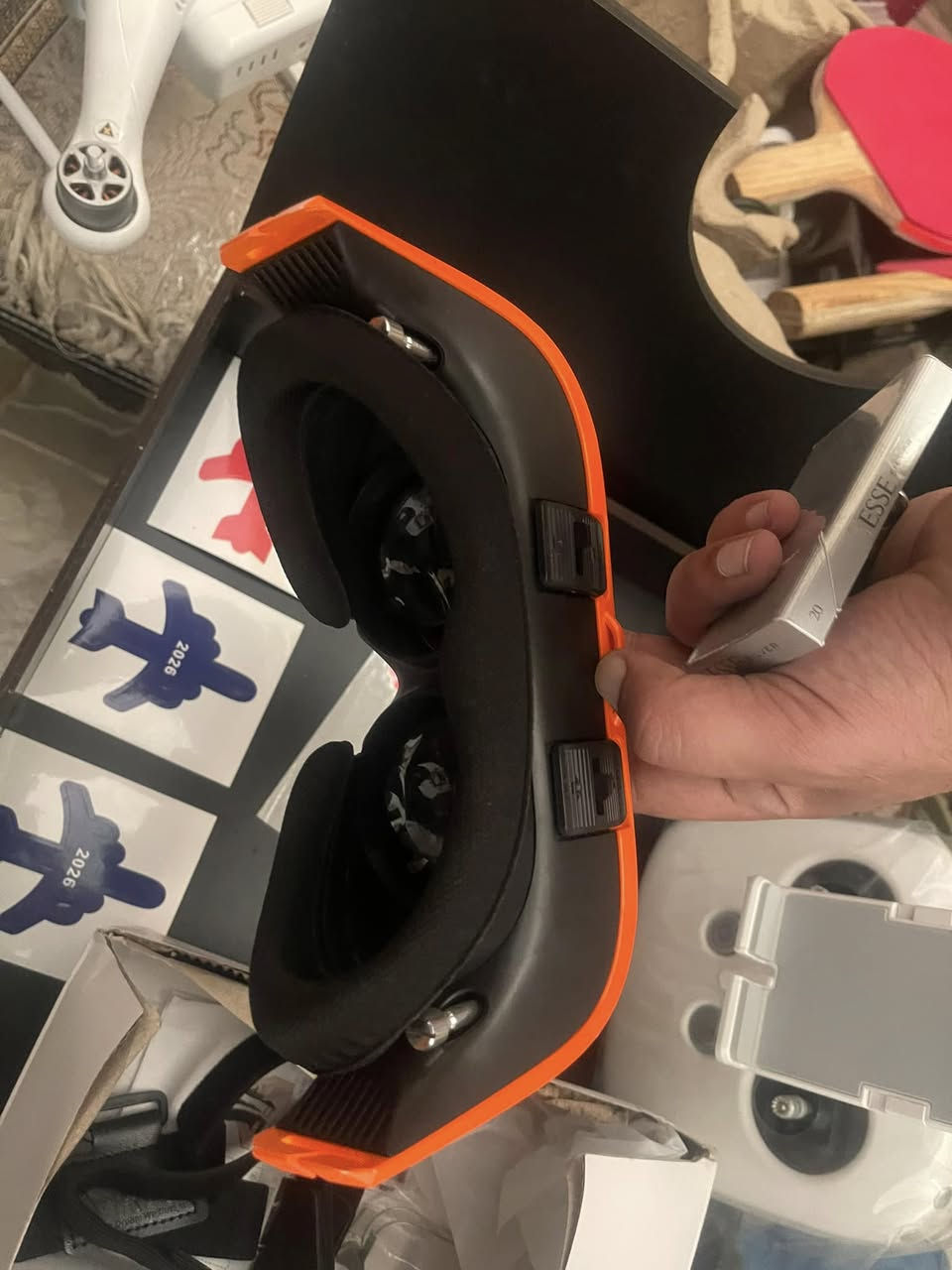 1. نظارة الطيران الافتراضي (FPV Goggles)
 الوصف: هي الأداة التي تحملها بيدك باللونين الأسود والبرتقالي.
 الوظيفة: تحتوي على شاشة وعدسات داخلية تعرض لك بثاً مباشراً وحياً من الكاميرا المثبتة على مقدمة الدرون، لتعيش تجربة الطيران وكأنك داخل مقصورة القيادة.
 الملحقات: يظهر أسفلها الحزام المطاطي الأسود (Headstrap) الخاص بتثبيت النظارة على الرأس داخل علبتها الكرتونية.


**إذا كنت صاحب هذا الإعلان وتريد حذفه لأي سبب، رجاءا أرسل رسالة إلى الدعم الفني**