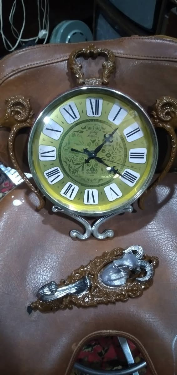 ساعة حائط مميزة جداً وتحمل طابعاً كلاسيكياً عتيقاً (Vintage/Retro)، وتحديداً من الساعات التي اشتهرت بها شركة "ريذم" (Rhythm) أو "كاسيو" (Casio) اليابانية في فترة السبعينيات والثمانينيات، والتي كانت تعتمد على تصاميم وزخارف بلاستيكية أو معدنية تحاكي الأنماط الأوروبية الكلاسيكية الكثيفة. مواصفات 
​ التصميم والهيكل الخارجي (The Aesthetic)
​ التصميم مستوحى من شكل "ساعة الجيب الكلاسيكية العتيقة" من حيث وجود الحلقة العلوية الكبيرة والمقبض، ولكن تمت إضافة عناصر زخرفية بارزة تمنحها طابعاً ملكياً أو ريفياً أوروبياً (Baroque / Rococo Style).
​التماثيل والزخارف: * رأس الكبش (أو الوعل): نلاحظ وجود رأس كبش بقرون دائرية مميزة في أعلى الساعة باللون الفضي المحاط بإطار بني مزخرف، وتتكرر رؤوس الكباش على الجانبين الأيمن والأيسر كحوامل أو مقابض جانبية. هذا الرمز غالباً ما يرمز للقوة أو يرتبط بتصاميم الساعات الجدارية المخصصة لغرف الضيوف أو المكاتب الفخمة.
​القاعدة السفلية: مزينة بزخرفة معدنية ملتوية باللون الفضي (Scrollwork) لتعطي توازناً بصرياً مع الجزء العلوي.
 الميناء والعقارب (The Dial & Hands)
​الخلفية (الميناء): تأتي باللون الأخضر الزيتوني المتدرج إلى الأصفر (Sunburst/Gradient Effect)، وهو لون كلاسيكي شهير جداً في حقبة السبعينيات. يوجد في منتصف الميناء نقوش وزخارف نباتية ناعمة ملتفة (Filigree Pattern).
​الأرقام: أرقام رومانية كبيرة باللون الأسود موضوعة داخل مربعات أو مخروطيات بيضاء بارزة، مما يسهل قراءتها ويمنح الميناء بُعداً ثلاثي الأبعاد.
​العقارب: العقارب مصممة على الطراز الكلاسيكي (Retro/Ornate Hands) مع وجود عقرب ثوانٍ نحيف.
​الكتابة: يظهر في النصف السفلي من الميناء كلمة "QUARTZ"، مما يؤكد أن الساعة تعمل بحركة الكوارتز الحديثة (التي تعتمد على البطارية) وليست حركة ميكانيكية بالزنبرك، وهو ما كان ثورة في دقة الوقت خلال تلك الحقبة.
 التحليل الفني والمنشأ
​الحركة الداخلية: تعمل الساعة بنظام الكوارتز (Battery-powered Quartz Movement)، وغالباً ما تكون الماكينة الداخلية يابانية الصنع (مثل ماكينات Rhythm الموثوقة جداً في امتصاص الطاقة وتعمير البطارية).
​المواد المستخدمة: الهيكل الخارجي مصنع من البلاستيك المقوى عالي الجودة (Hard Injection-Molded Plastic) الذي كان يُدهن بطبقات تحاكي الخشب الداكن أو النحاس والمعدن الفضي، وهي تقنية برعت فيها المصانع اليابانية لإنتاج ساعات حائط خفيفة الوزن ذات مظهر فخم وثقيل.


**إذا كنت صاحب هذا الإعلان وتريد حذفه لأي سبب، رجاءا أرسل رسالة إلى الدعم الفني**