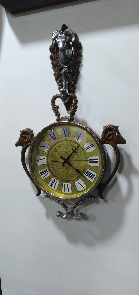ساعة حائط مميزة جداً وتحمل طابعاً كلاسيكياً عتيقاً (Vintage/Retro)، وتحديداً من الساعات التي اشتهرت بها شركة "ريذم" (Rhythm) أو "كاسيو" (Casio) اليابانية في فترة السبعينيات والثمانينيات، والتي كانت تعتمد على تصاميم وزخارف بلاستيكية أو معدنية تحاكي الأنماط الأوروبية الكلاسيكية الكثيفة. مواصفات 
​ التصميم والهيكل الخارجي (The Aesthetic)
​ التصميم مستوحى من شكل "ساعة الجيب الكلاسيكية العتيقة" من حيث وجود الحلقة العلوية الكبيرة والمقبض، ولكن تمت إضافة عناصر زخرفية بارزة تمنحها طابعاً ملكياً أو ريفياً أوروبياً (Baroque / Rococo Style).
​التماثيل والزخارف: * رأس الكبش (أو الوعل): نلاحظ وجود رأس كبش بقرون دائرية مميزة في أعلى الساعة باللون الفضي المحاط بإطار بني مزخرف، وتتكرر رؤوس الكباش على الجانبين الأيمن والأيسر كحوامل أو مقابض جانبية. هذا الرمز غالباً ما يرمز للقوة أو يرتبط بتصاميم الساعات الجدارية المخصصة لغرف الضيوف أو المكاتب الفخمة.
​القاعدة السفلية: مزينة بزخرفة معدنية ملتوية باللون الفضي (Scrollwork) لتعطي توازناً بصرياً مع الجزء العلوي.
 الميناء والعقارب (The Dial & Hands)
​الخلفية (الميناء): تأتي باللون الأخضر الزيتوني المتدرج إلى الأصفر (Sunburst/Gradient Effect)، وهو لون كلاسيكي شهير جداً في حقبة السبعينيات. يوجد في منتصف الميناء نقوش وزخارف نباتية ناعمة ملتفة (Filigree Pattern).
​الأرقام: أرقام رومانية كبيرة باللون الأسود موضوعة داخل مربعات أو مخروطيات بيضاء بارزة، مما يسهل قراءتها ويمنح الميناء بُعداً ثلاثي الأبعاد.
​العقارب: العقارب مصممة على الطراز الكلاسيكي (Retro/Ornate Hands) مع وجود عقرب ثوانٍ نحيف.
​الكتابة: يظهر في النصف السفلي من الميناء كلمة "QUARTZ"، مما يؤكد أن الساعة تعمل بحركة الكوارتز الحديثة (التي تعتمد على البطارية) وليست حركة ميكانيكية بالزنبرك، وهو ما كان ثورة في دقة الوقت خلال تلك الحقبة.
 التحليل الفني والمنشأ
​الحركة الداخلية: تعمل الساعة بنظام الكوارتز (Battery-powered Quartz Movement)، وغالباً ما تكون الماكينة الداخلية يابانية الصنع (مثل ماكينات Rhythm الموثوقة جداً في امتصاص الطاقة وتعمير البطارية).
​المواد المستخدمة: الهيكل الخارجي مصنع من البلاستيك المقوى عالي الجودة (Hard Injection-Molded Plastic) الذي كان يُدهن بطبقات تحاكي الخشب الداكن أو النحاس والمعدن الفضي، وهي تقنية برعت فيها المصانع اليابانية لإنتاج ساعات حائط خفيفة الوزن ذات مظهر فخم وثقيل.


**إذا كنت صاحب هذا الإعلان وتريد حذفه لأي سبب، رجاءا أرسل رسالة إلى الدعم الفني**