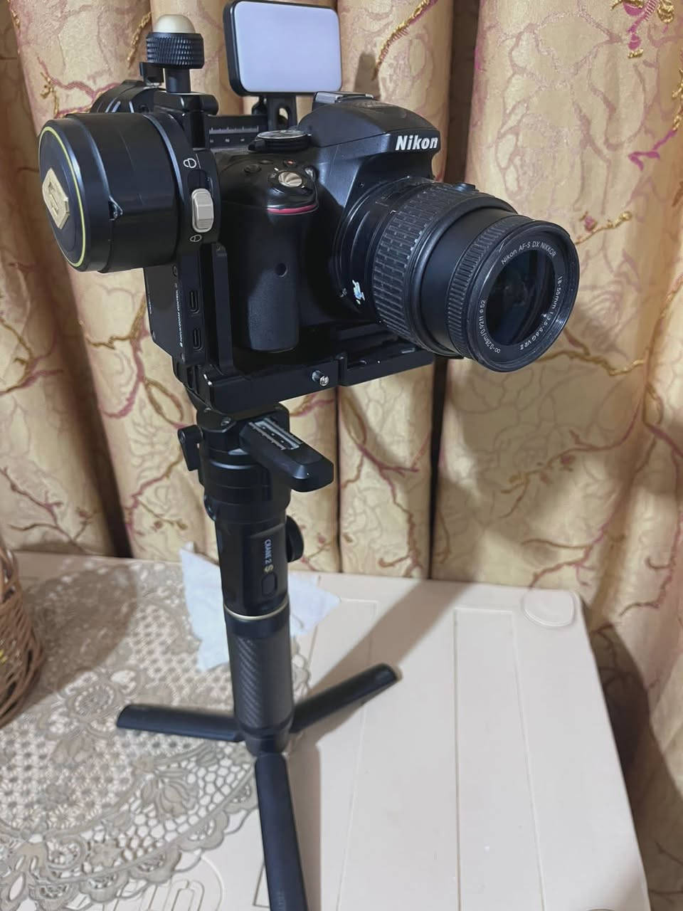 مكلف بل نشر
1. الكاميرا والعدسة (Body & Lens)
 نوع الكاميرا: Nikon D5300 (كاميرا احترافية من فئة DSLR معروفة جداً بموثوقيتها).
 دقة وميزات الكاميرا:
 مستشعر بدقة 24.2 ميجابكسل يعطي صوراً عالية الدقة وتفاصيل حادة.
 شاشة خلفية متحركة (تفتح وتفتر) وهي ممتازة جداً لتصوير الفيديو والـ Vlogs أو السيلفي.
 دعم تصوير فيديو بدقة Full HD (1080p) بسرعة تصل إلى 60 إطار في الثانية، مما يعطي سلاومة ونعومة بالفيديو.
 تحتوي على Wi-Fi مدمج لنقل الصور مباشرة للموبايل.
 العدسة (Lens): عدسة نيكون الأساسية AF-S NIKKOR 18-55mm f/3.5-5.6G VR.
 ميزة الزووم: مثل ما ذكرتِ، الصورة تتكبر وتتصغر (من 18mm للقطات الواسعة إلى 55mm للقطات القريبة والبورتريه).
 مانع اهتزاز (VR): العدسة مجهزة بنظام تقليل الاهتزاز الاهتزاز البصري لضمان صور واضحة بدون غواش حتى أثناء الحركة.
2. مانع الاهتزاز الاحترافي (Gimbal)
 النوع الموديل: Zhiyun Crane 2S (واضح من الشعار المكتوب باللون الذهبي والكتابة على يد المانع).
 وظيفة الجهاز: هذا الجهاز مخصص للمصورين وصناع المحتوى المحترفين؛ حيث يوازن الكاميرا تماماً ويمنع أي اهتزاز أثناء المشي أو الركض، ليظهر الفيديو ناعماً وسلساً مثل تصوير الأفلام السينمائية.
 المميزات الظاهرة بالصور:
 يد كربون فايبر (Carbon Fiber) خفيفة وقوية.
 أقفال أمان للمحاور (Axis Locks) تسهل عملية الموازنة والتنقل (كما في ⁠image.png⁠ و ⁠image_6.png⁠).
 قاعدة ترايبود (Tripod) سفلية صغيرة لثبات المانع على الأسطح.
3. الملحقات الإضافية (Accessories)
 إضاءة LED مدمجة: مثبتة أعلى مانع الاهتزاز (تظهر بوضوح في ⁠image_2.png⁠ و ⁠image_4.png⁠)؛ توفر إضاءة مستمرة وممتازة لتصوير الوجوه أو الفيديوهات في الأماكن المظلمة.
 حقيبة الحمل (Case): تظهر جزئياً في الخلفية بـ ⁠image_10.png⁠ وهي الحقيبة الأصلية لمانع الاهتزاز لحمايته أثناء النقل ***********
