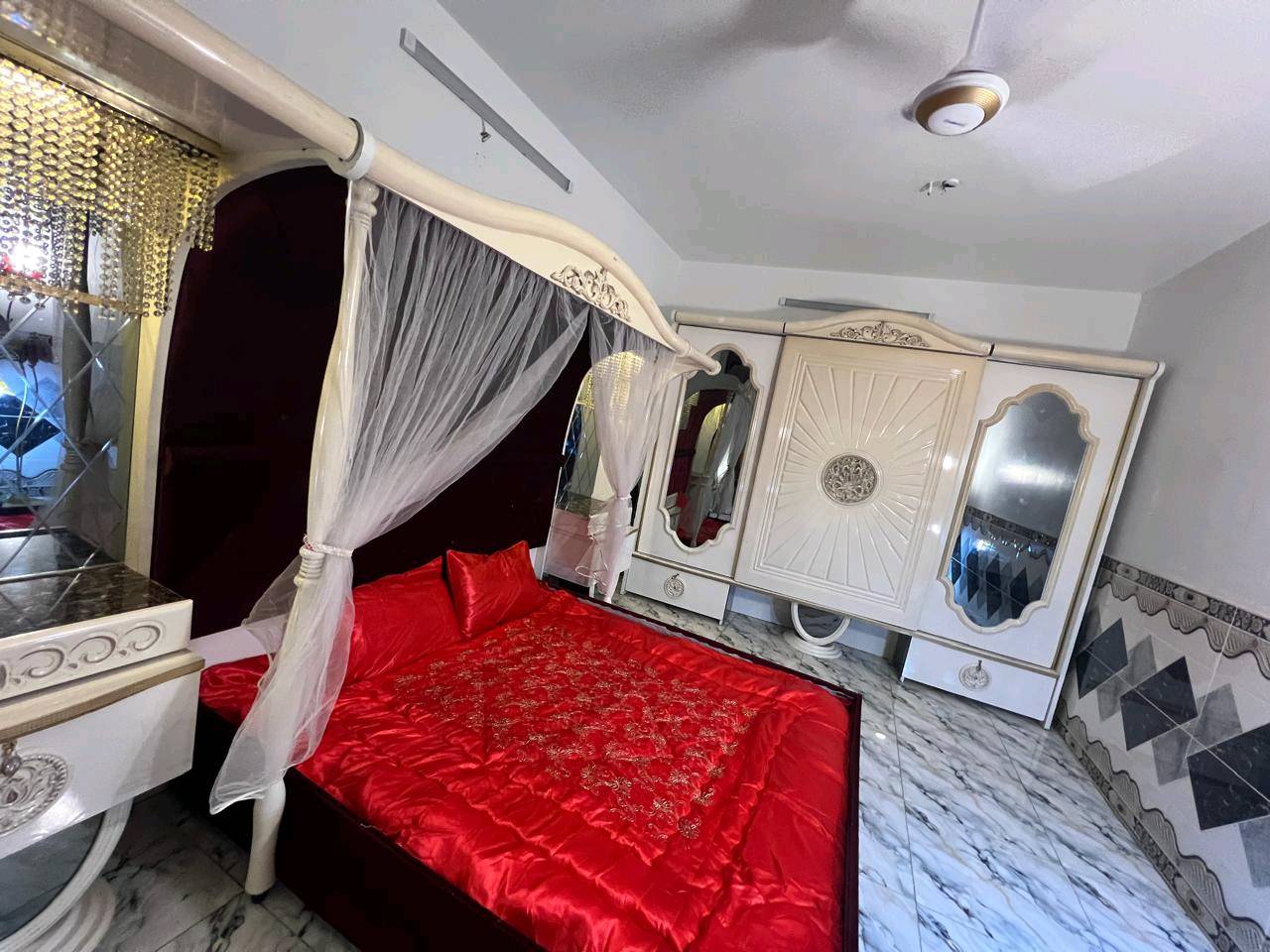 غرفة نوم تركية بتصميم فاخر وجميل، تتكون من سرير مزود بستارة شبكية وستائر مضاءة، بالإضافة إلى خزانة كبيرة مع مرايا. التصميم يجمع بين اللونين الأبيض والذهبي مع تغطية السرير بلون أحمر فاقع، مناسب لإضافة لمسة من الأناقة والرفاهية لغرفتك.