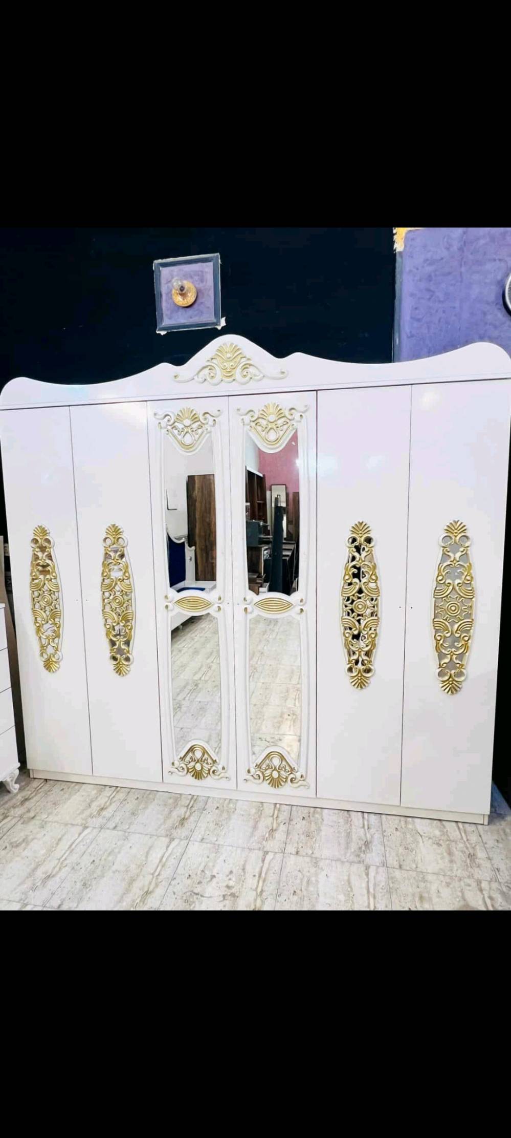 غرفة نوم فاخرة بتصميم كلاسيكي تشمل سرير مريح مع لوح أمامي مبطن باللون الأزرق الداكن وخزانة ملابس كبيرة مع مرايا زخرفية. تتميز الغرفة بتفاصيل ذهبية على الأبواب والأدراج، مما يضفي لمسة من الفخامة والرقي. الطقم يشمل أيضًا تسريحة مع مرآة كبيرة وكرسي مبطن لتوفير الراحة أثناء التحضير اليومي. الأثاث مصنوع بجودة عالية ويضفي لمسة فنية على أي غرفة نوم.