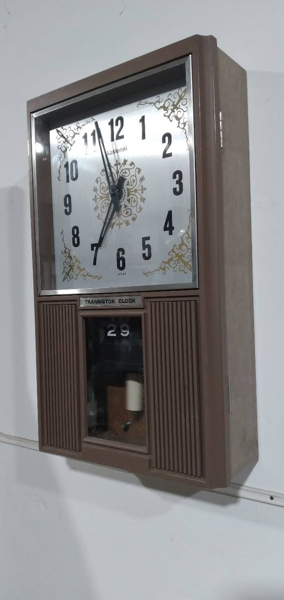 ***********
كلاسيكية رائعة جداً!  ساعة حائط يابانية عتيقة (Vintage) من إنتاج شركة National (ناشيونال - المعروفة حالياً باسم باناسونيك Panasonic)، وتحديداً من فئة ساعات "Transistor Clock" (ساعة ترانزستور) التي كانت شائعة جداً في فترة أواخر الستينيات والسبعينيات من القرن الماضي.
​الهيكل والتصميم الخارجي
​النمط المستوحى من الخمسينيات والستينيات (Mid-Century Modern): تتميز الساعة بهيكل مستطيل رأسي، مطلي باللون البني الكلاسيكي مع وجود خطوط رأسية (مشرشرة) على الجانبين السفليين تشبه فتحات المكبرات الصوتية في الراديو القديم، وهو أسلوب تصميمي كان دارجاً جداً ليعطي انطباعاً بالفخامة والدفء المنزلي.
​مينا الساعة (Dial): المينا يأتي بخلفية معدنية فضية لامعة (Brushed aluminum)، محاطة بزخارف ذهبية كلاسيكية في الأركان وفي المنتصف. الأرقام العربية كبيرة، سوداء، وواضحة جداً بخط سميك يسهل قراءته من مسافة بعيدة.
​العلامة التجارية والمنشأ:   شعار National أسفل الرقم 12، وفي الأسفل تماماً تحت الرقم 6 كُتبت عبارة JAPAN لتؤكد جودة الصناعة اليابانية في تلك الحقبة.
​ آلية العمل (الحركة والتكنولوجيا)
​Transistor Clock (ساعة الترانزستور): اللوحة الفضية الصغيرة أسفل المينا تحمل عبارة "TRANSISTOR CLOCK". هذه التكنولوجيا كانت طفرة في وقتها؛ فهي تمثل مرحلة انتقالية بين الساعات الميكانيكية بالكامل (التي تعتمد على الزنبرك والتعبئة اليدوية) وساعات الكوارتز الحديثة. تعمل هذه الساعة بالبطارية، حيث يقوم ترانزستور صغير بتنظيم النبضات الكهربائية لتحريك بندول مغناطيسي أو عجلة توازن، مما يحافظ على دقة الوقت دون الحاجة لتعبئتها يدوياً.
​البندول (Pendulum): في النافذة السفلية المحاطة بالإطار البني، يمكن رؤية جزء من البندول الداخلي الذي يتحرك يميناً ويساراً، وهو مغطى بزجاج شفاف يتيح رؤية الحركة الميكانيكية التي تعطي الساعة روحاً وجمالية خاصة.
​ الميزات الإضافية (التقويم)
​نافذة التاريخ واليوم (Day/Date Calendar): بجانب البندول في القسم السفلي، تحتوي الساعة على تقويم ميكانيكي يظهر اليوم والشهر (يظهر في الصورة رقم 29 واليوم "Tue" أي الثلاثاء). هذا النوع من التقويم يتغير تلقائياً مع حركة عقارب الساعة عند منتصف الليل.
​باختصار، هذه الساعة ليست مجرد أداة لمعرفة الوقت، بل هي قطعة فنية تجسد ذروة الصناعة الهندسية والجمالية اليابانية في السبعينيات، وتعتبر اليوم من القطع المميزة جداً لهواة جمع المقتنيات والأنتيكات الإلكترو-ميكانيكية.
